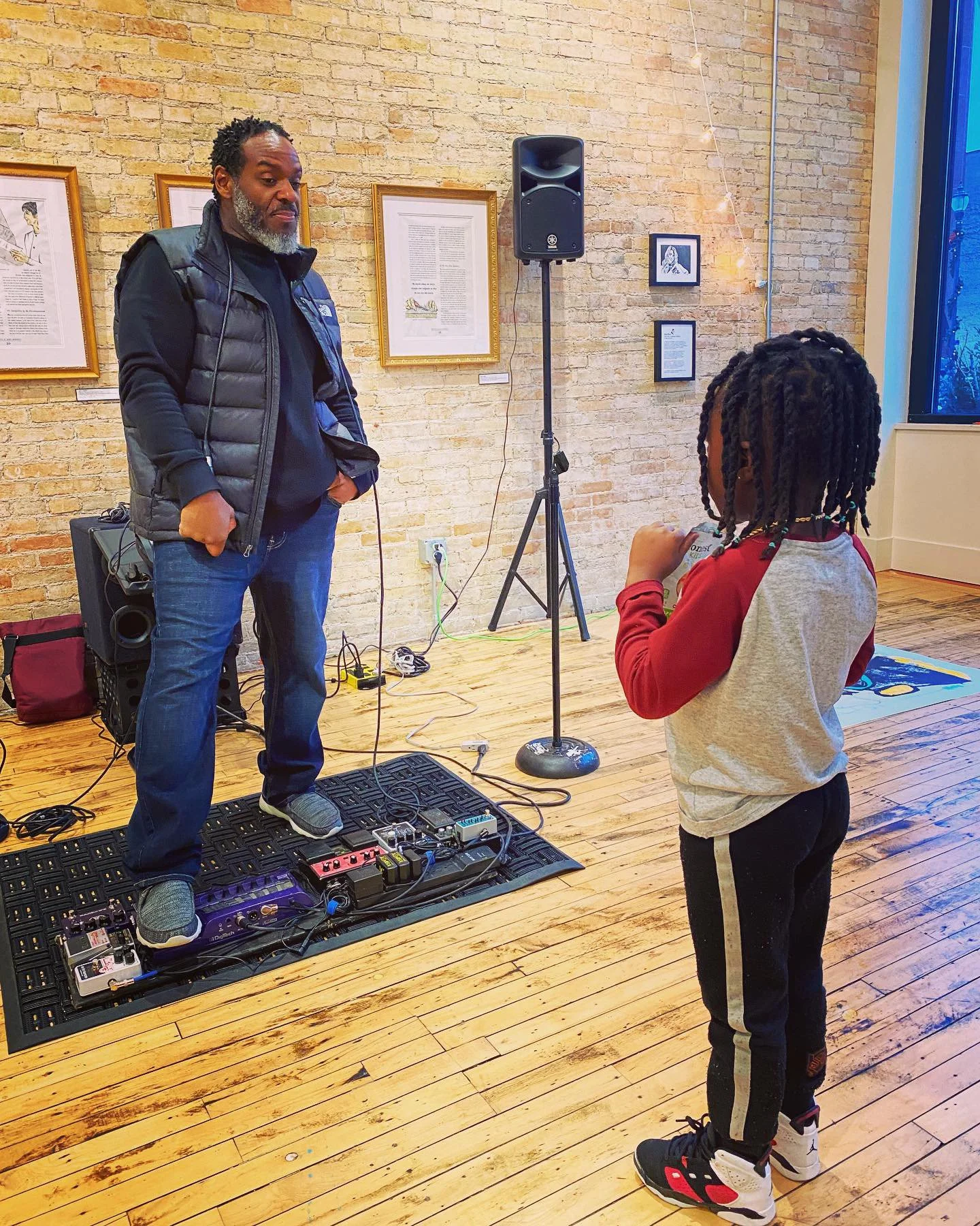 A man with gray beard and black vest is standing next to a young boy with braided hair holding a card, in an art gallery or studio with a brick wall and framed artwork behind them. The man is near music equipment on the floor.