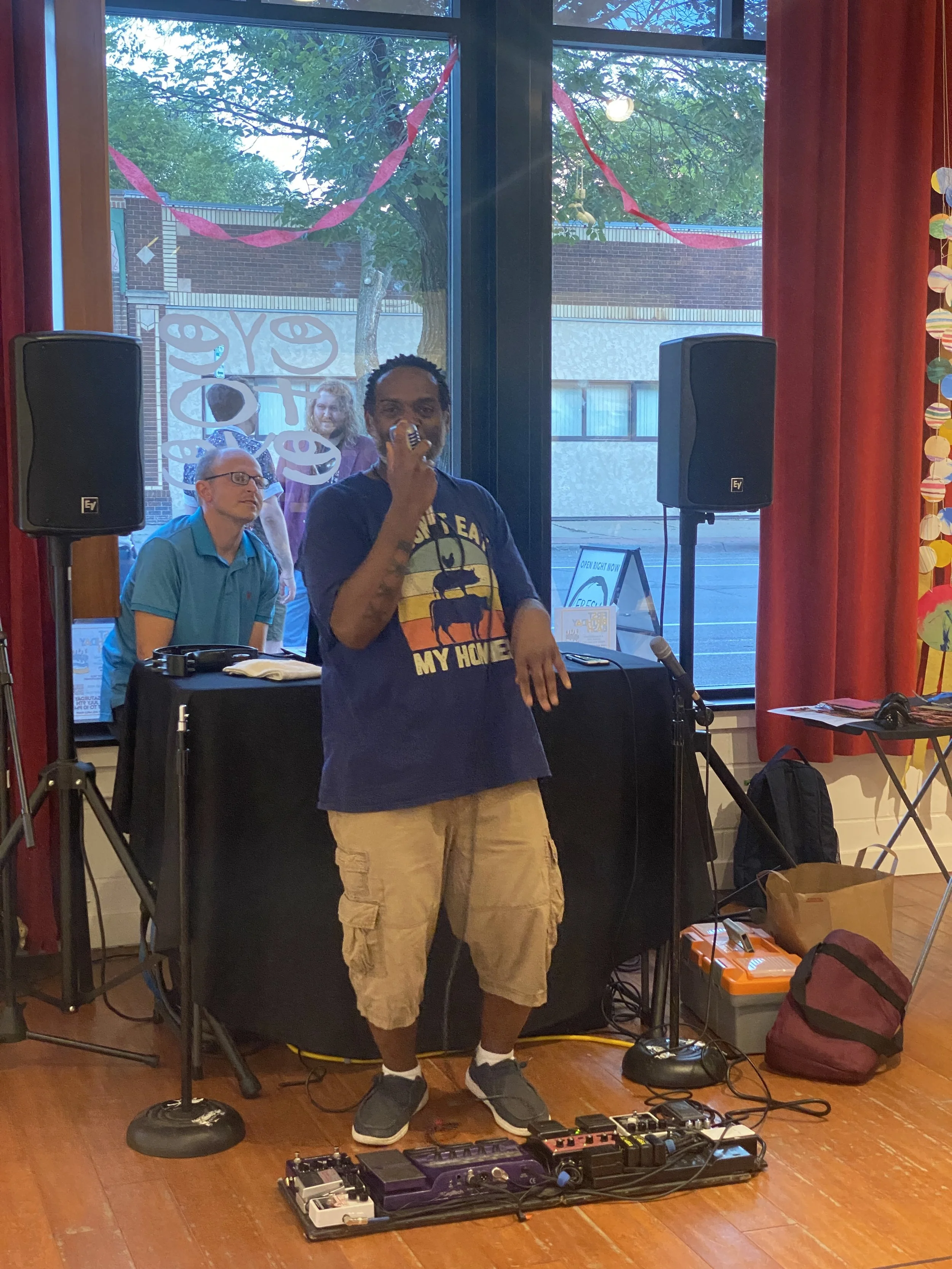 A man in a dark T-shirt and khaki shorts stands on a wooden floor, speaking into a microphone, with musical equipment and foot pedals at his feet. Behind him, there are two large black speakers, a table with a DJ setup, and two people in the backgrou