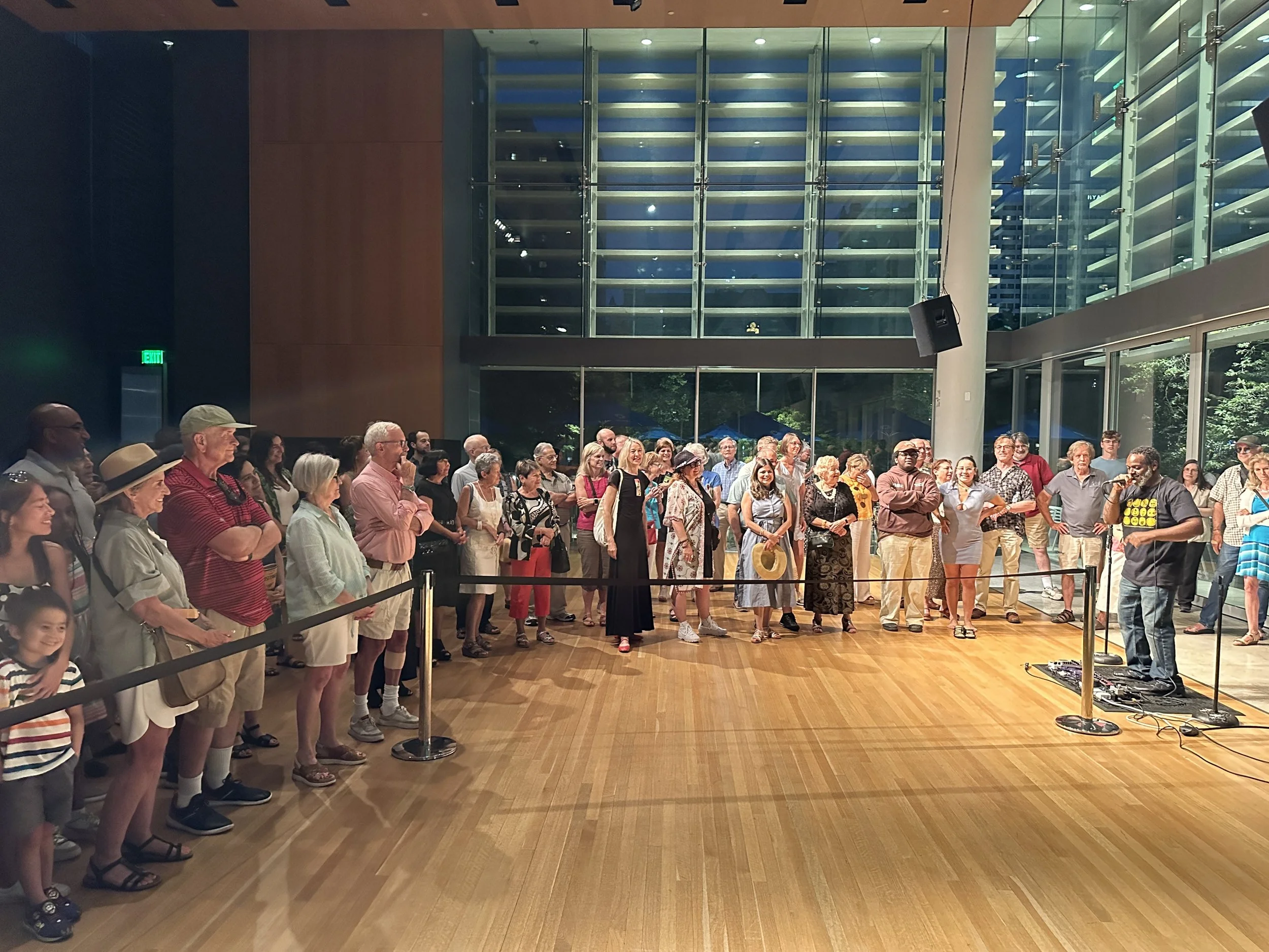 Group of people attending an indoor event or presentation, standing in a line, facing to the right. The room has large windows and wooden flooring, with some individuals smiling and taking photos.