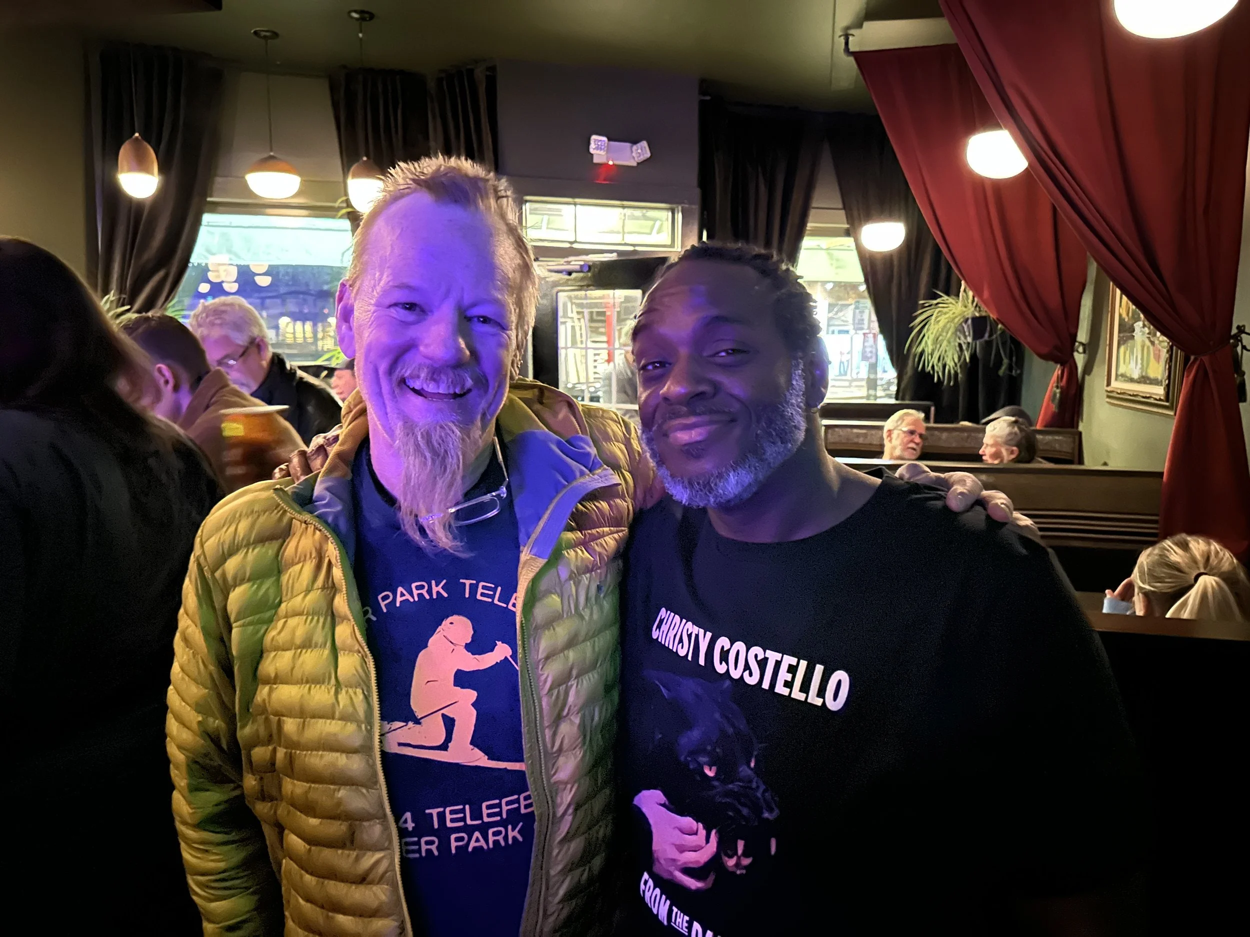 Two men smiling at the camera inside a dimly lit restaurant or bar. One man on the left has a red beard and is wearing a yellow jacket and a black T-shirt. The other man on the right has short curly hair and is wearing a black T-shirt. In the backgro