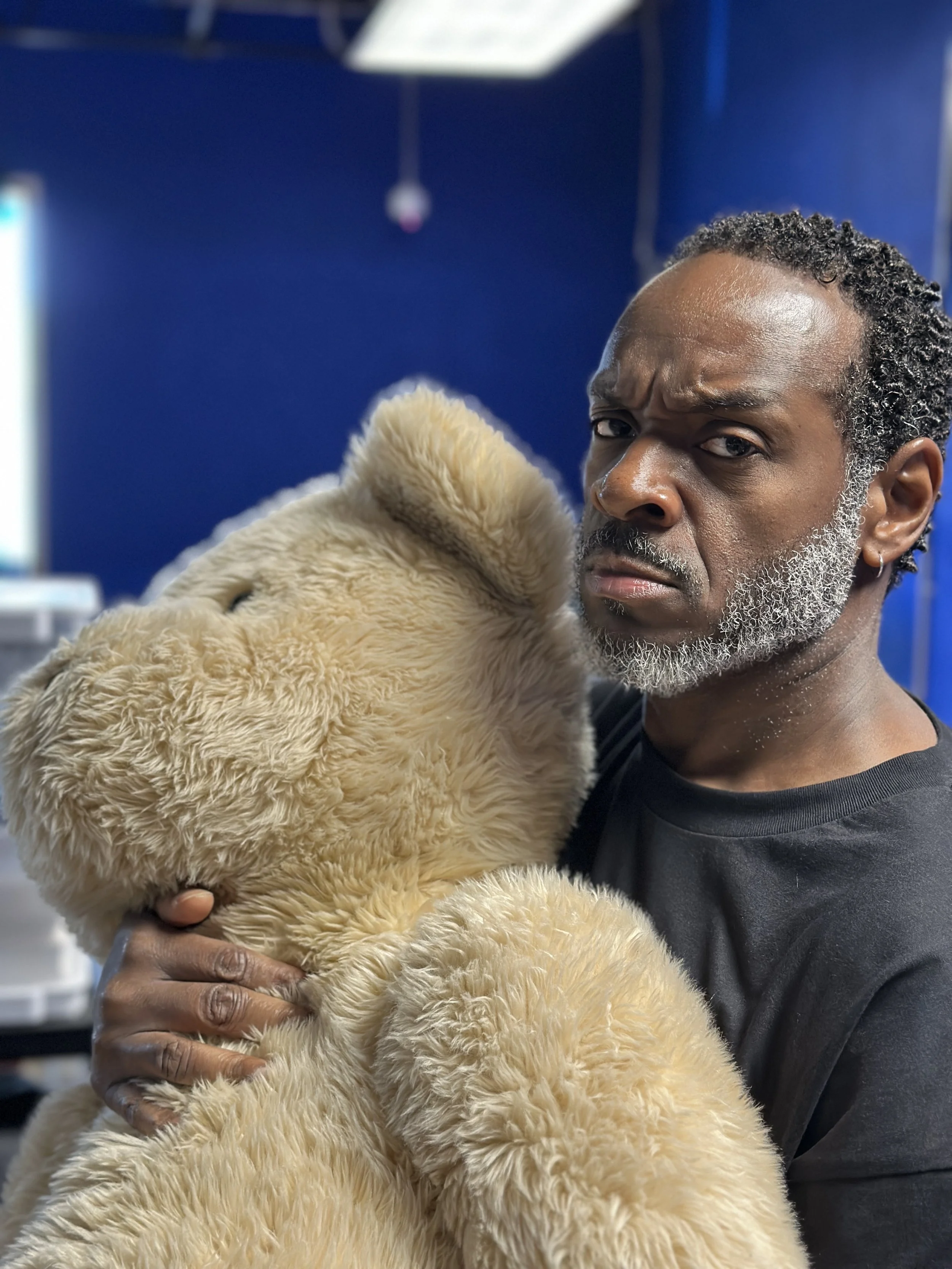 A man holding a fluffy beige teddy bear, with a serious expression, in front of a blue wall.