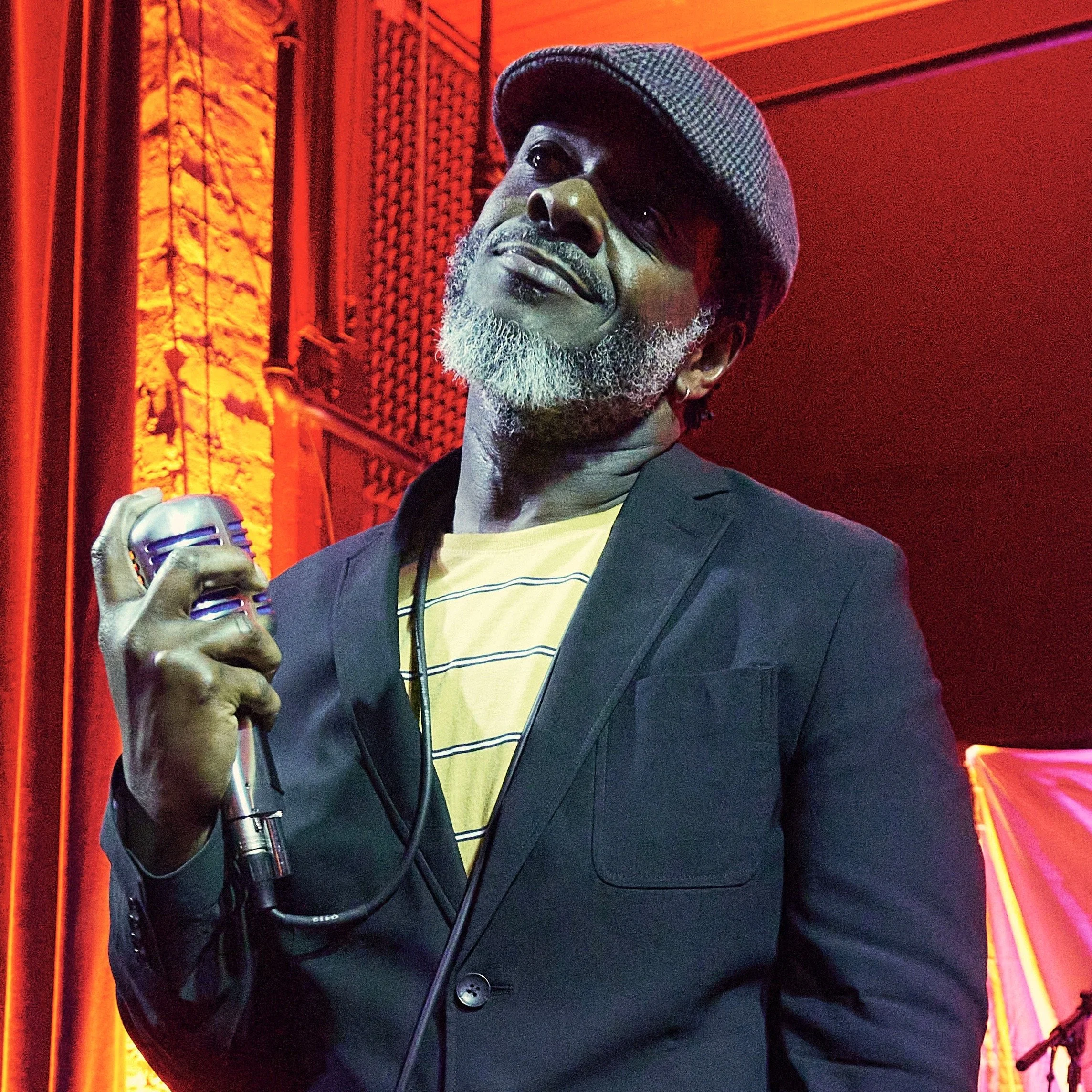 A man with a gray beard wearing a checkered flat cap, black jacket, and yellow t-shirt singing into a microphone against a red background with yellow lighting accents.