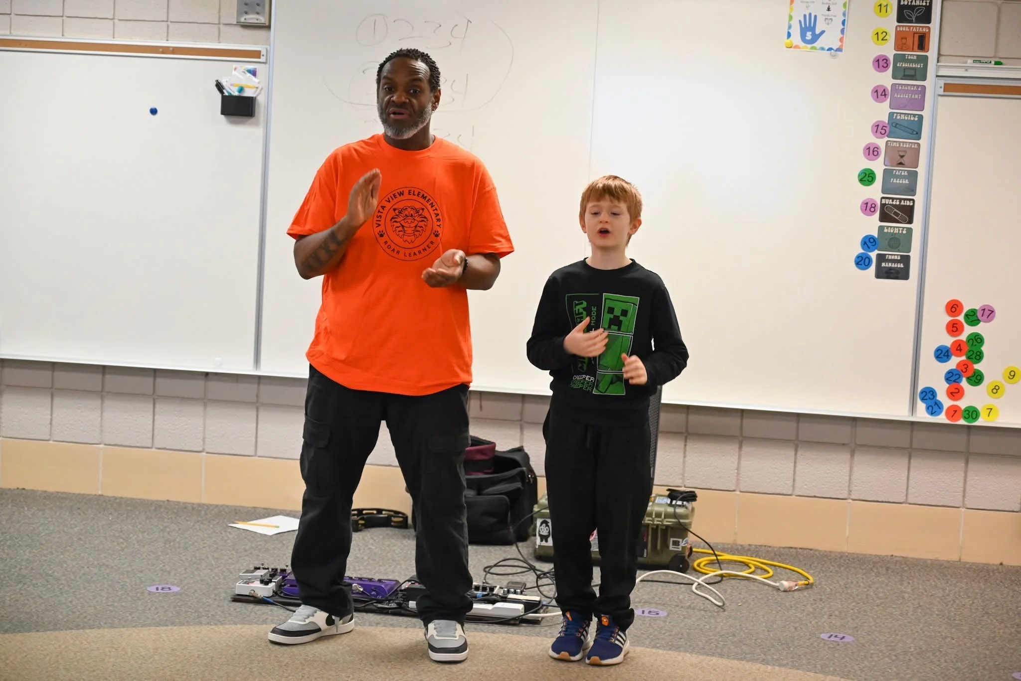 A man and a boy standing in a classroom, possibly giving a presentation, with musical equipment and education materials around them, in front of a whiteboard.