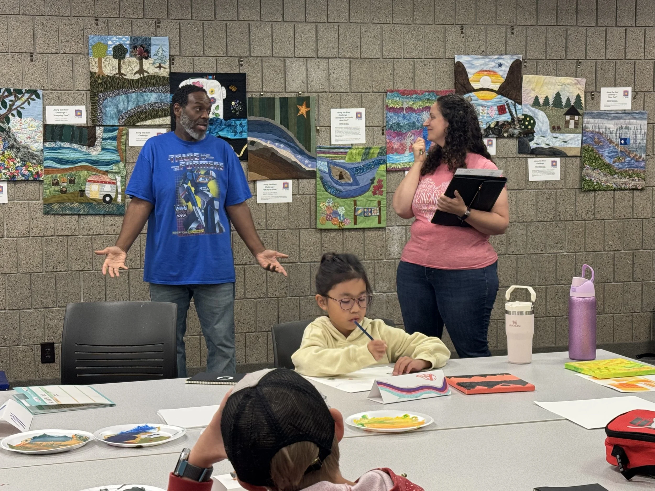 Two adults are standing and talking in front of a wall with colorful quilt art, while children sit at a table with art supplies and paint plates in the foreground.