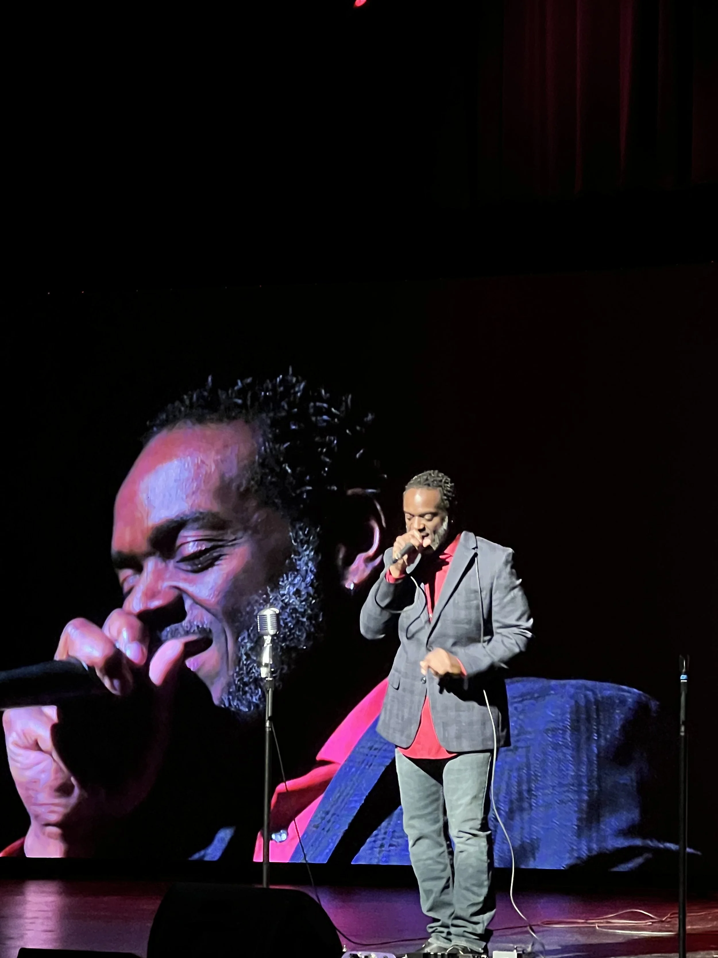 A male performer singing on stage, with a large screen behind displaying an enlarged face of himself, wearing a gray suit, red shirt, and gray pants, holding a microphone, and standing in front of a music stand on the stage.