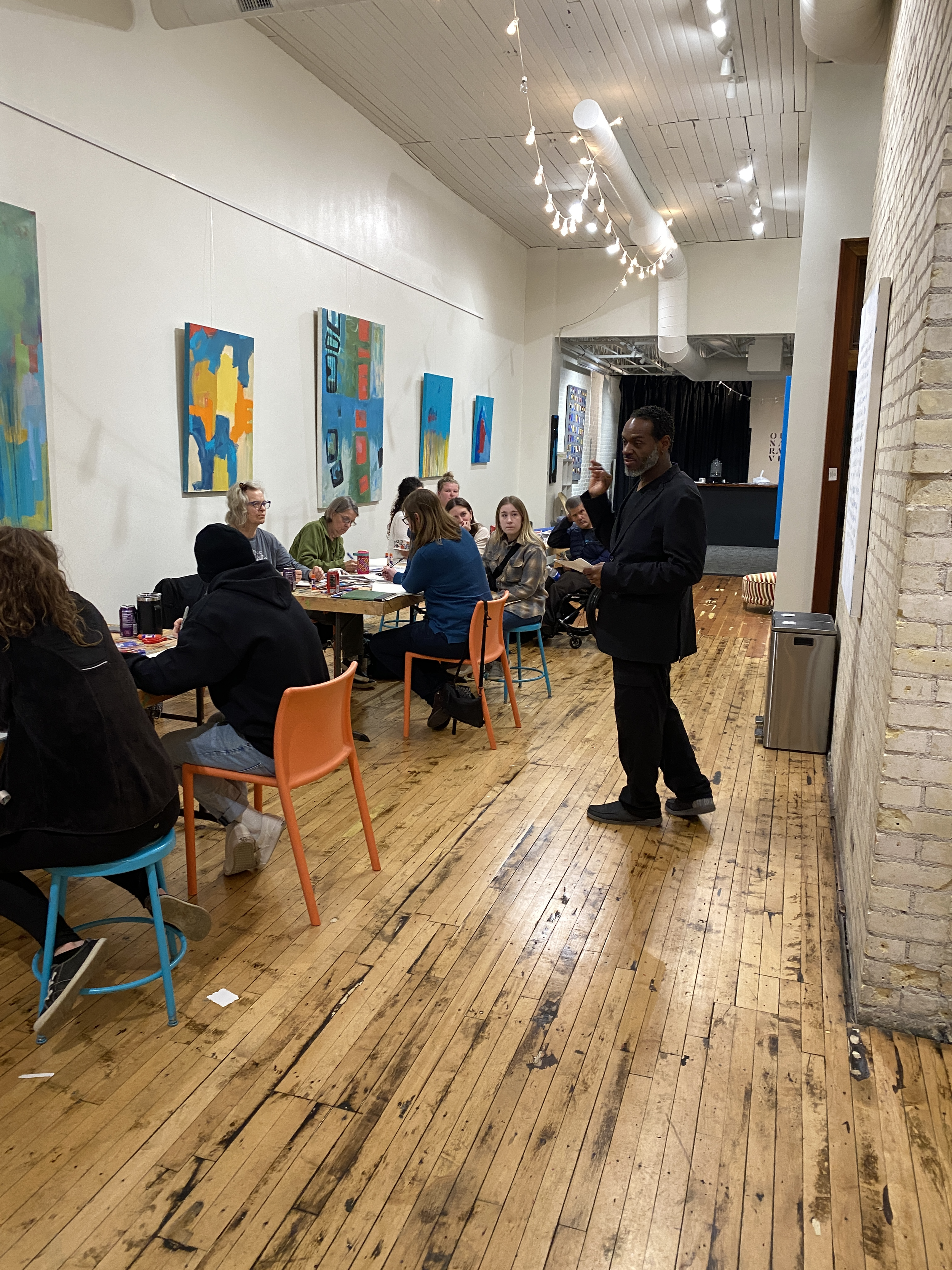 A man in a black suit speaking or presenting to a group of people seated at long tables in an art gallery or studio with colorful paintings on the walls and string lights hanging from the ceiling.