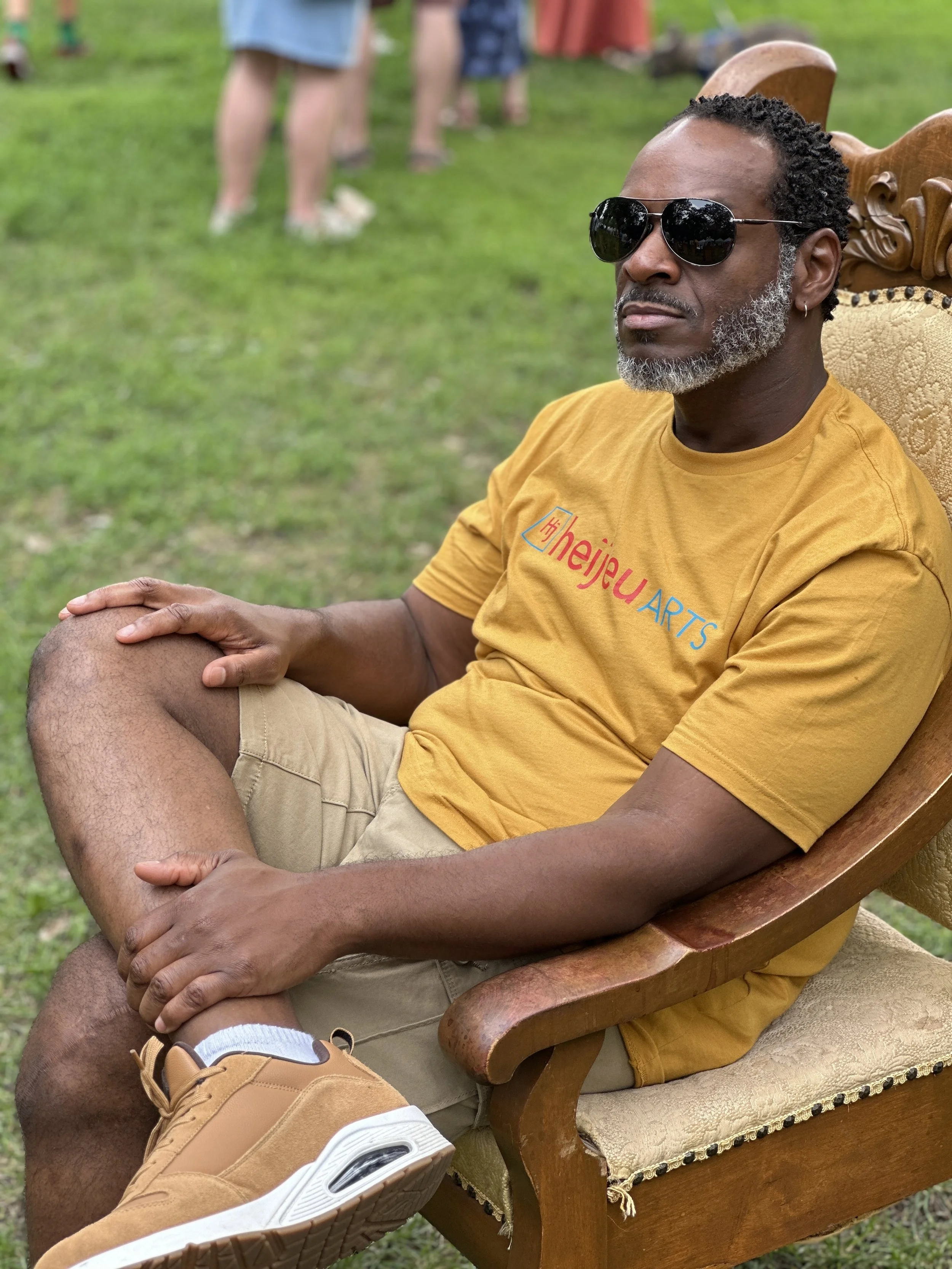 A man wearing sunglasses and a yellow T-shirt seated on a wooden chair outdoors, with his legs crossed and hand resting on his knee. The background shows blurred grass and people standing.