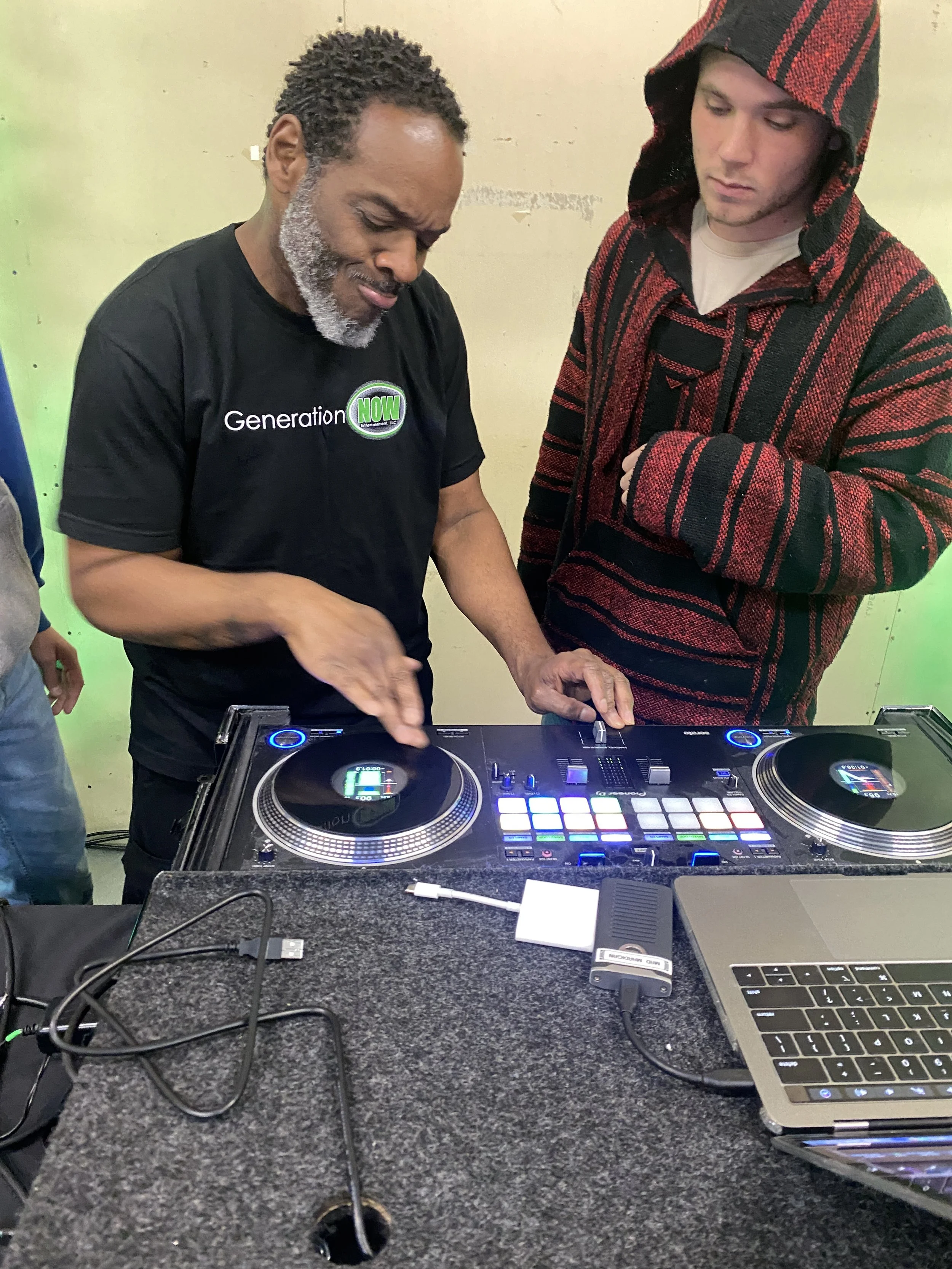 A man with gray beard and black t-shirt standing behind DJ equipment and mixing music with another young man wearing a hoodie. There is a laptop, DJ controller, and cables on a gray felt-covered table.