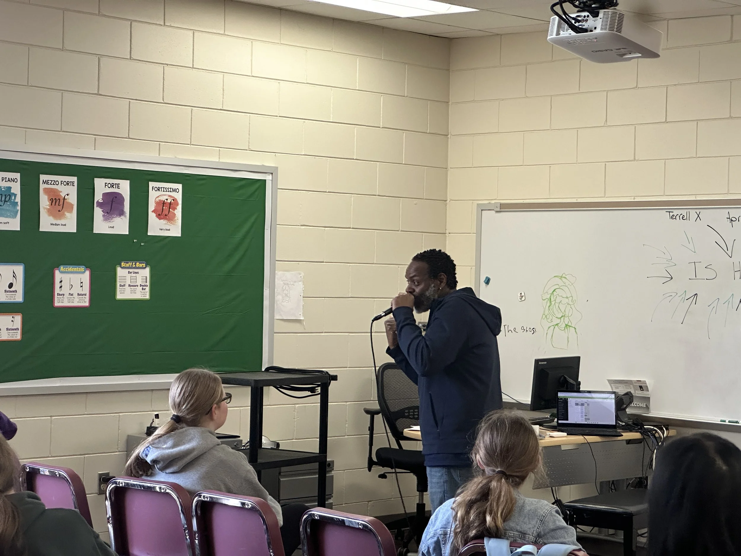 A classroom scene with students seated, a man standing near a whiteboard and speaking into a microphone, and a projector mounted on the ceiling. Several posters with musical dynamics are visible on a green bulletin board.