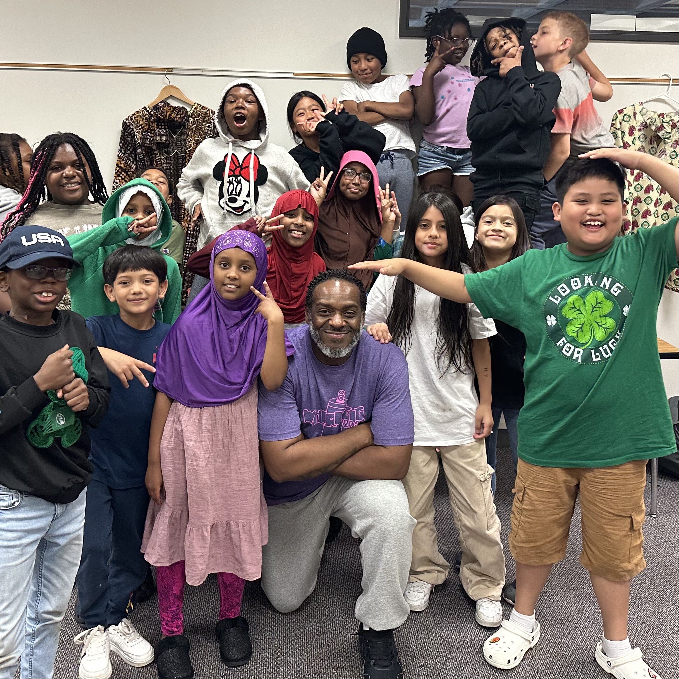 A diverse group of children and one adult woman posing together indoors, smiling and making various gestures. Some children are wearing colorful clothing, hoodies, and hats, while others are in casual attire. The setting appears to be a classroom or 