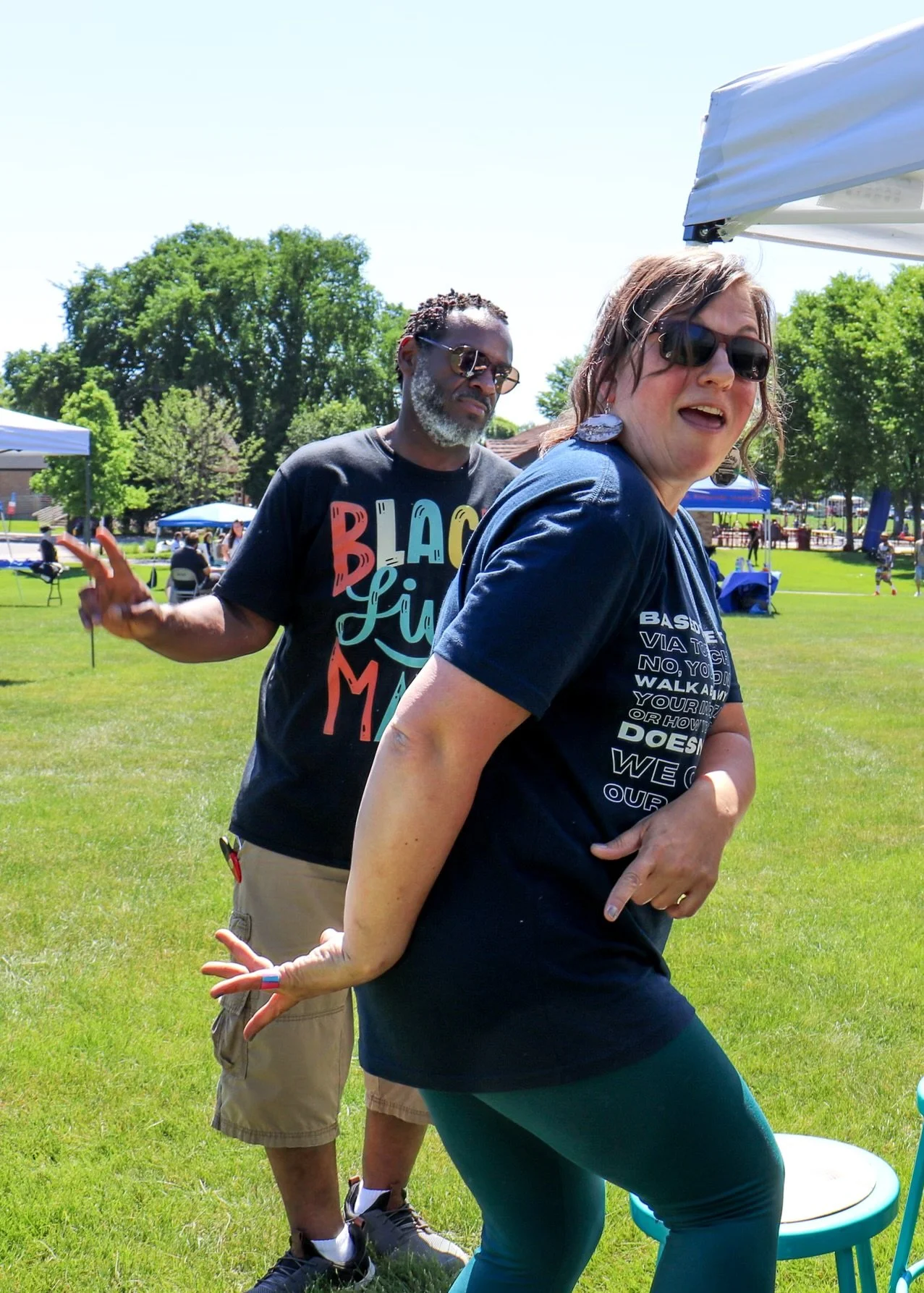 Two women and a man outdoors at a park, participating in a fun activity. The woman in front wears sunglasses, a dark T-shirt, and teal leggings. The man behind wears sunglasses, a black T-shirt with colorful text, and beige shorts. The woman in the b