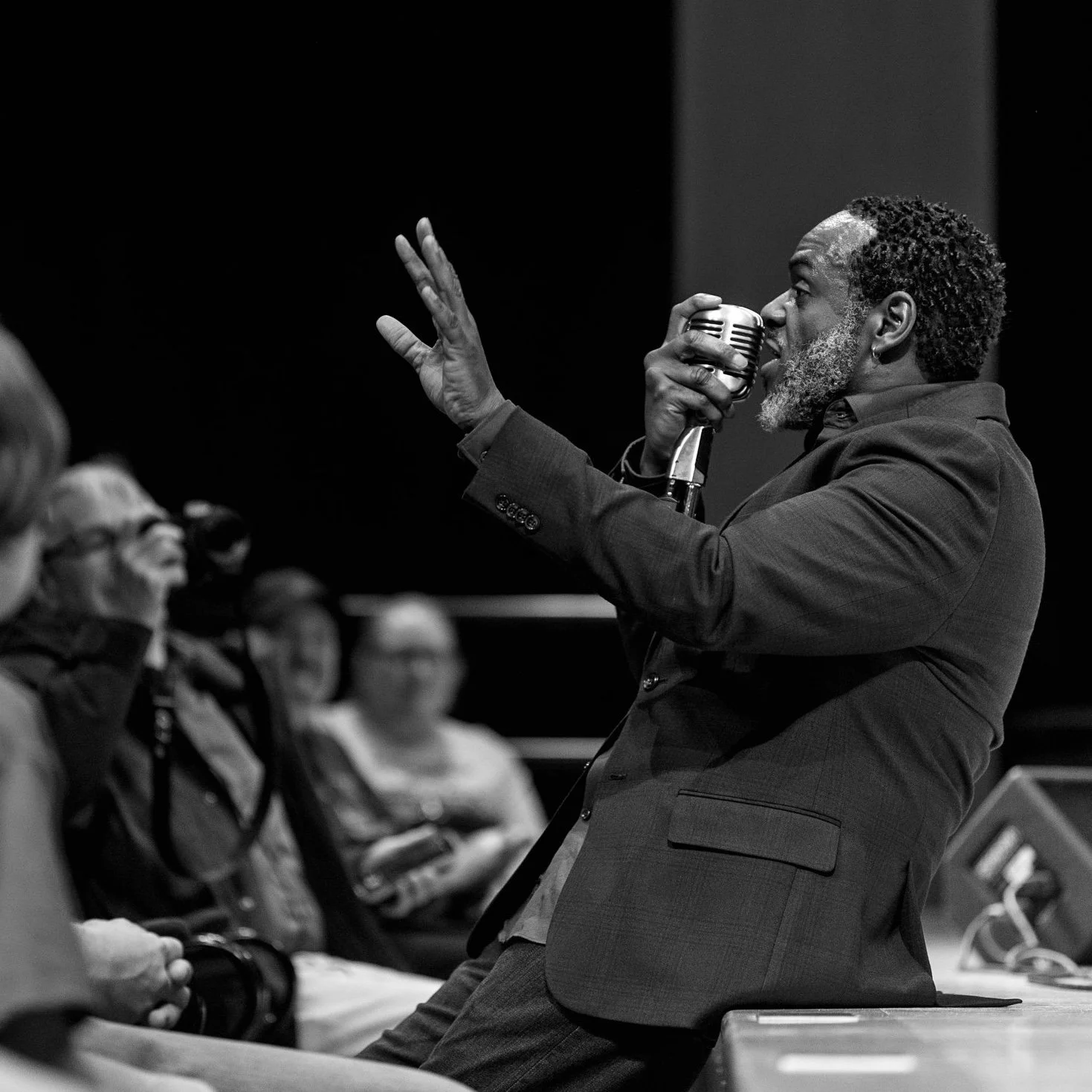 A man with curly hair and a beard speaking into a microphone during a presentation or speech, with an audience listening attentively.