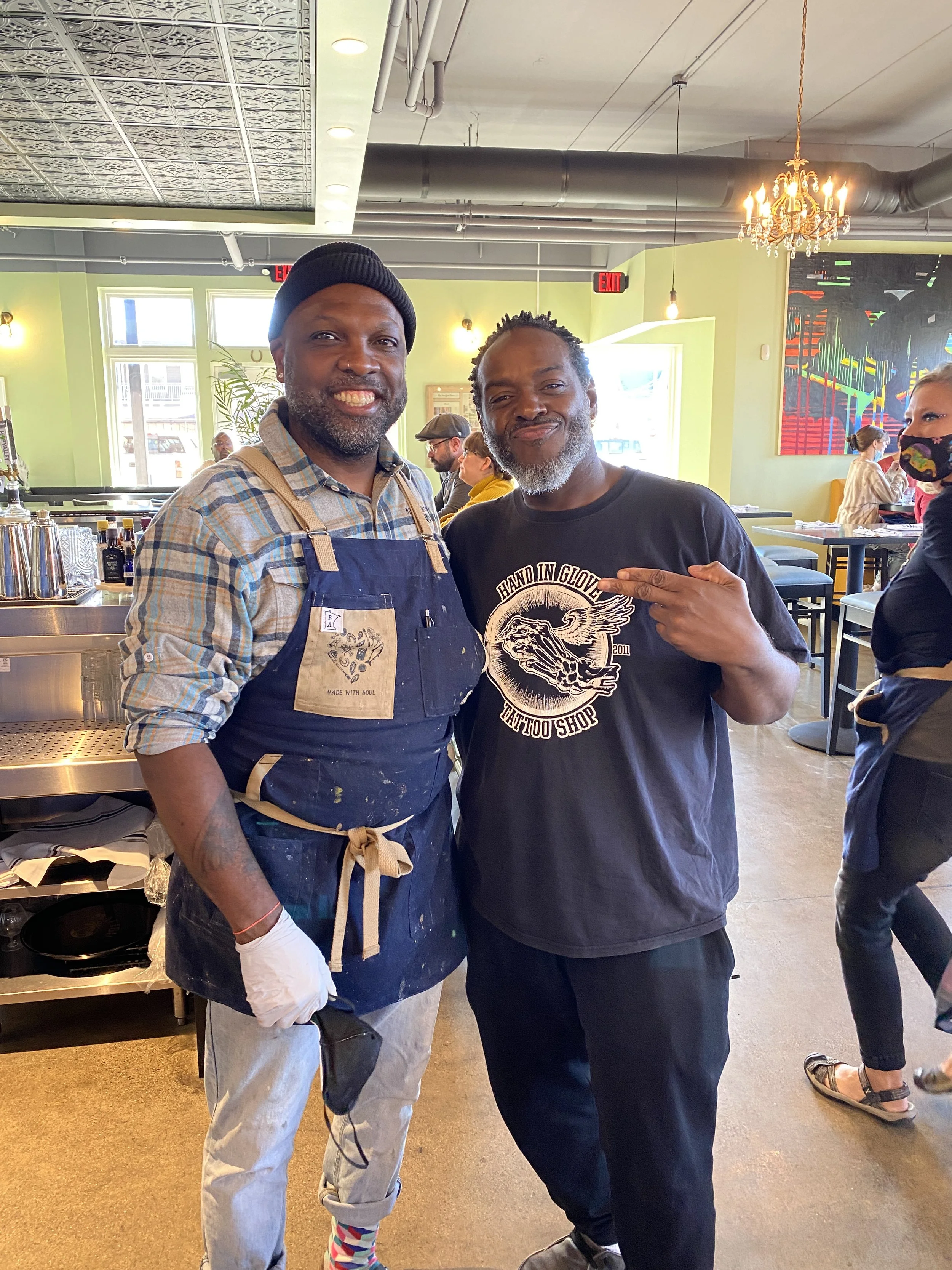 Two men standing in a restaurant, smiling at the camera. The man on the left is wearing a black beanie, a plaid shirt, an apron, gray jeans with rolled-up cuffs, and colorful socks. The man on the right has a gray beard, is wearing a black T-shirt wi