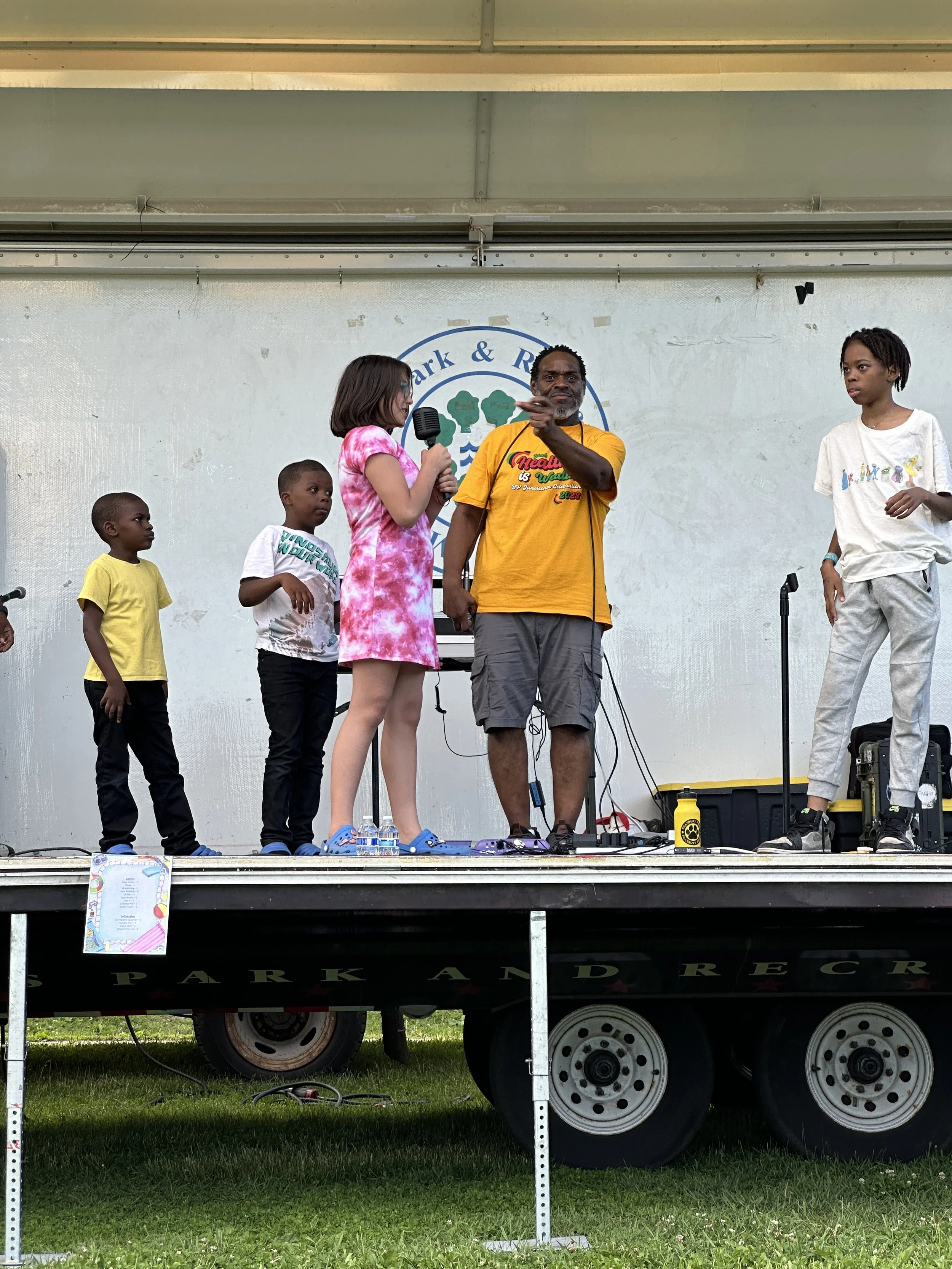 A group of six people on a stage, four children and two adults, during an outdoor event. The children are standing on the left and right, with the woman microphone in her hand and the man pointing at her. The stage has a banner with a park logo behin