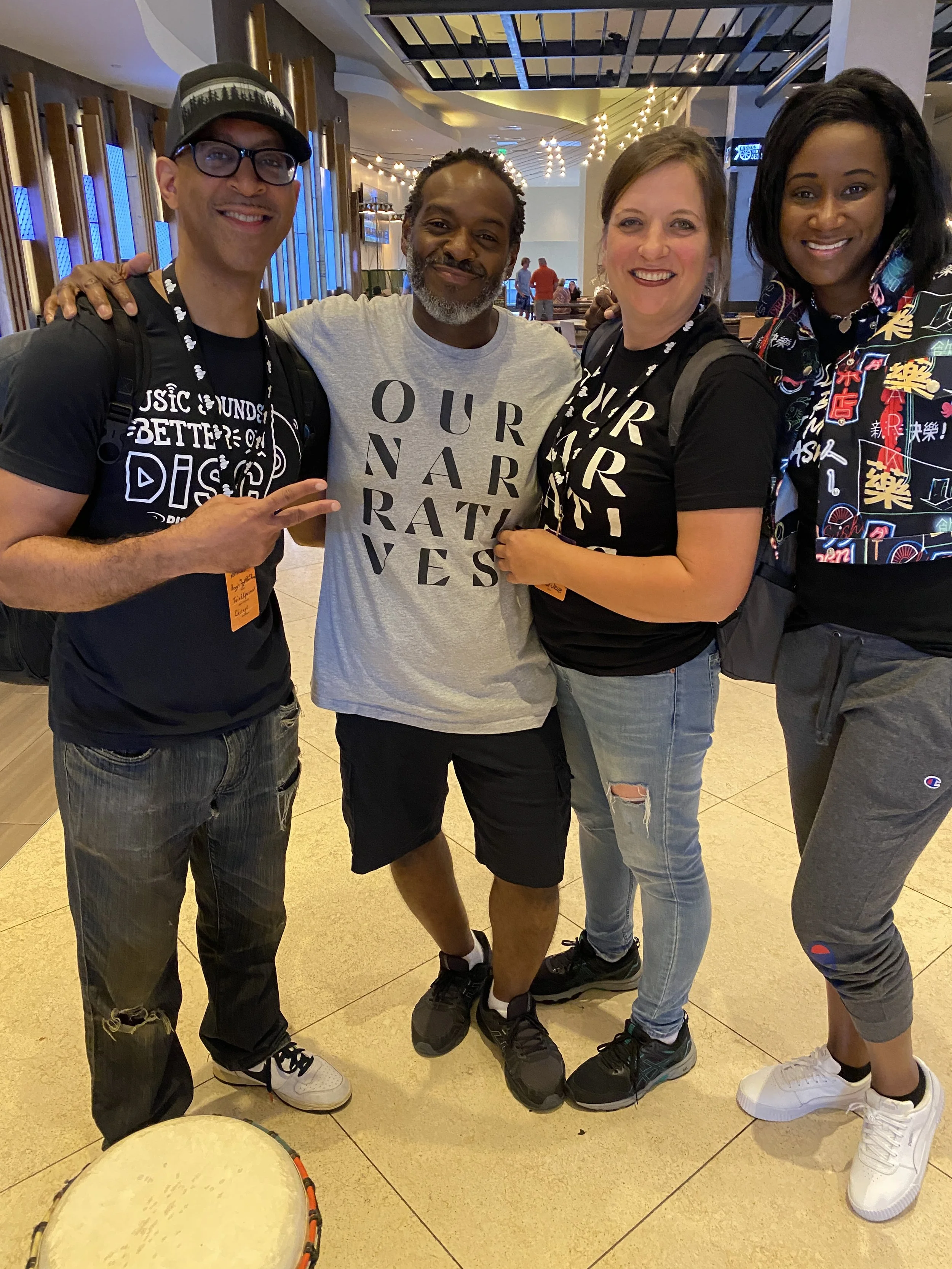 Four people standing inside a modern, well-lit building, smiling and posing for a photo. Two of them are wearing black t-shirts with text, one is wearing a grey t-shirt, and the fourth is wearing a colorful shirt with Asian characters. There is a dru