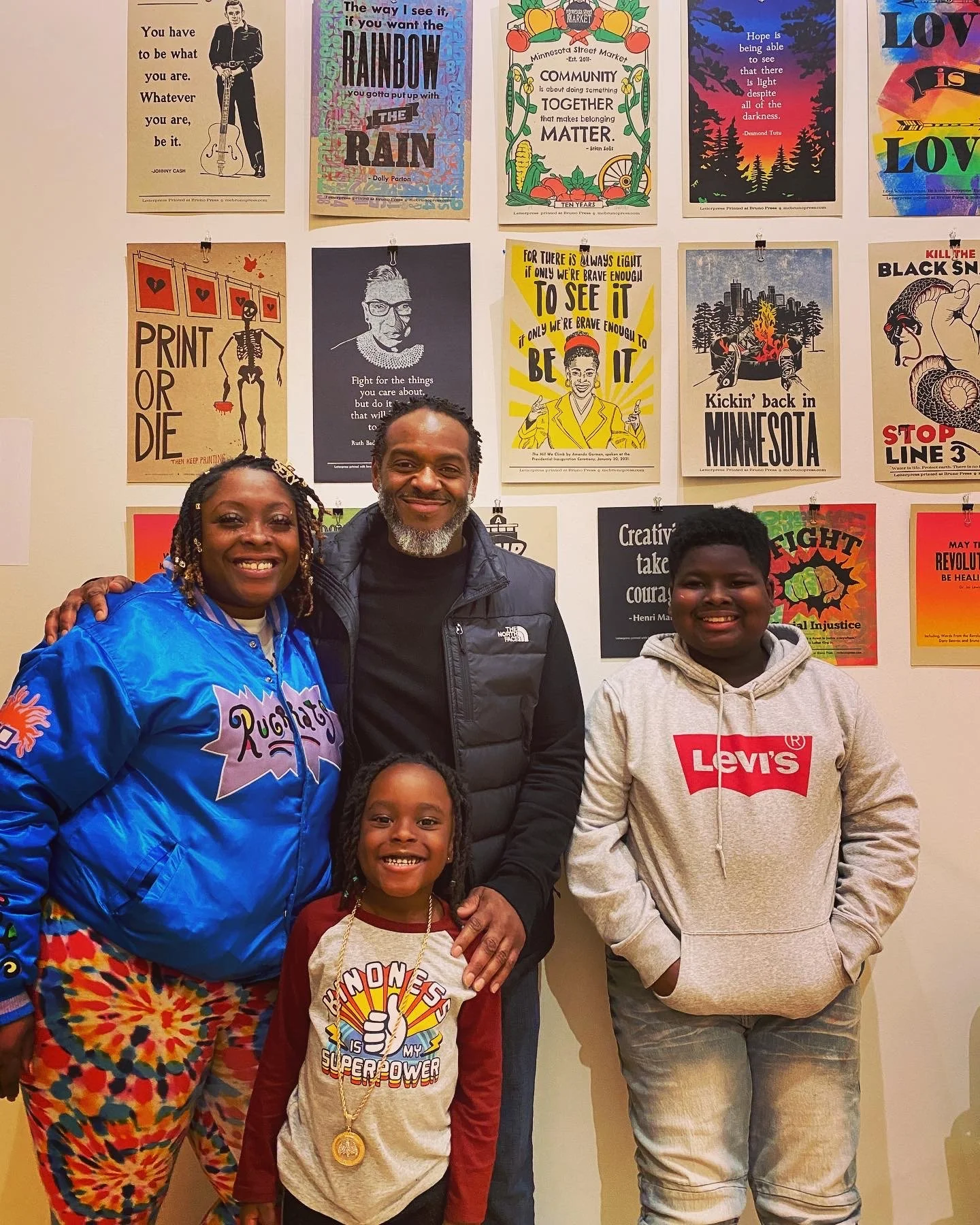 A group of five people, three children and two adults, standing in front of a wall covered with colorful posters with inspirational quotes and artwork. The people are smiling and appear happy.