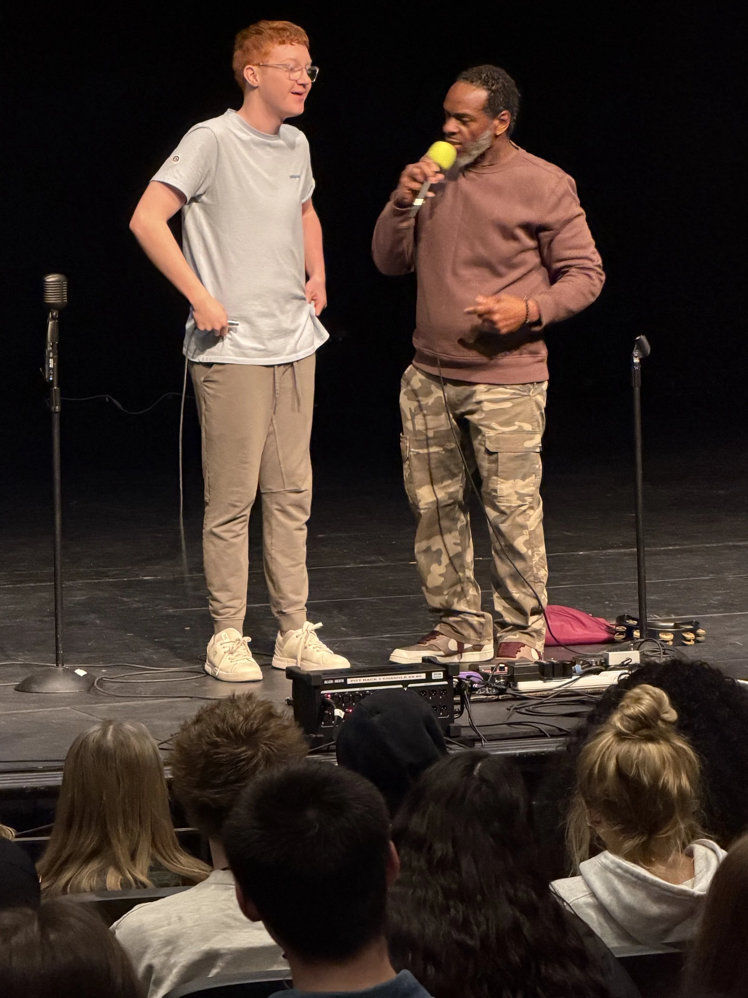 Two men stand on a stage, one holding a microphone and speaking, the other holding an object in his hand. They are surrounded by microphones and audio equipment. An audience watches in the foreground.