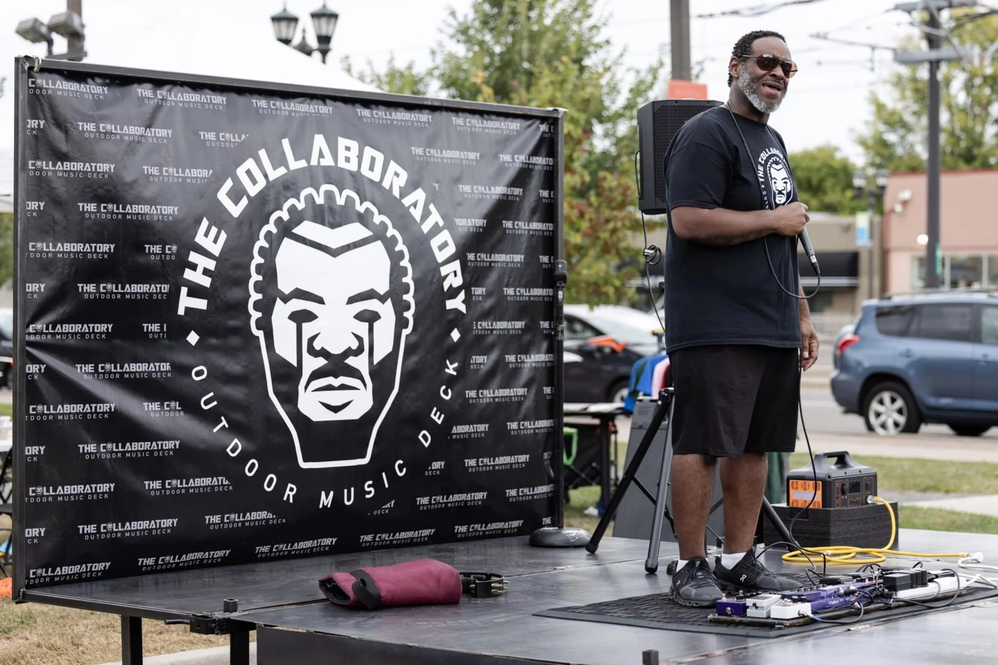 A man holding a microphone stands beside a speaker and music equipment during an outdoor event. Behind him is a black banner with a stress face logo and text that reads 'The Collaborative Outdoor Music Deck.'