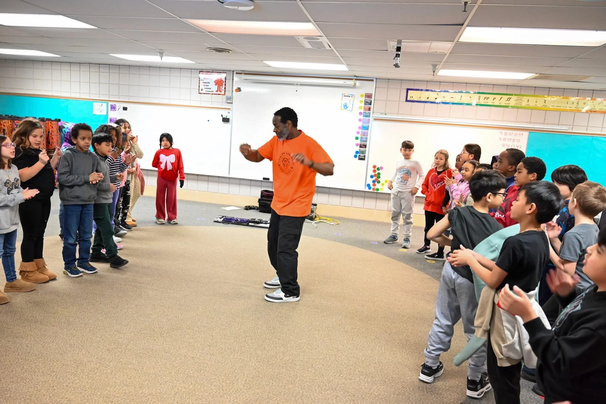 A man, possibly a music teacher or performer, leads a group of young students in a classroom, engaging them in an activity involving clapping or movement. The classroom has whiteboard, colorful decorations, and educational posters.