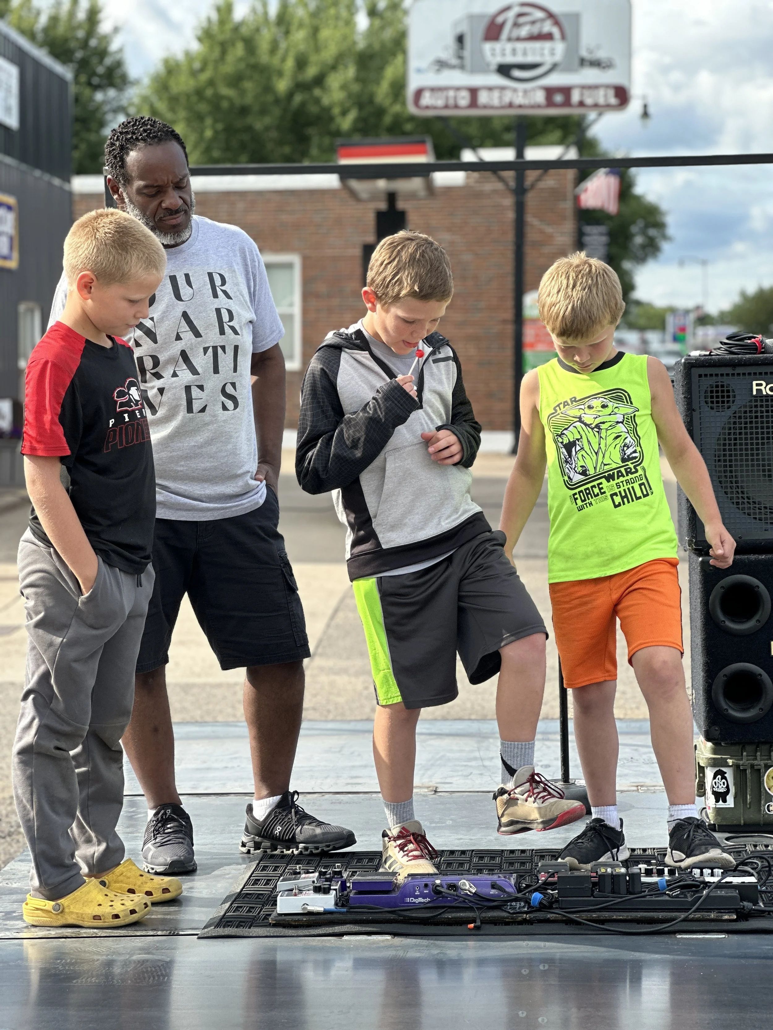 A manstands next to three boys on a stage. The boys are looking down at music or equipment, one in a red and black t-shirt, another in a gray hoodie, and the third in a bright green star wars t-shirt. There are speakers and audio equipment on the stage, and outdoor trees and a building are visible in the background.