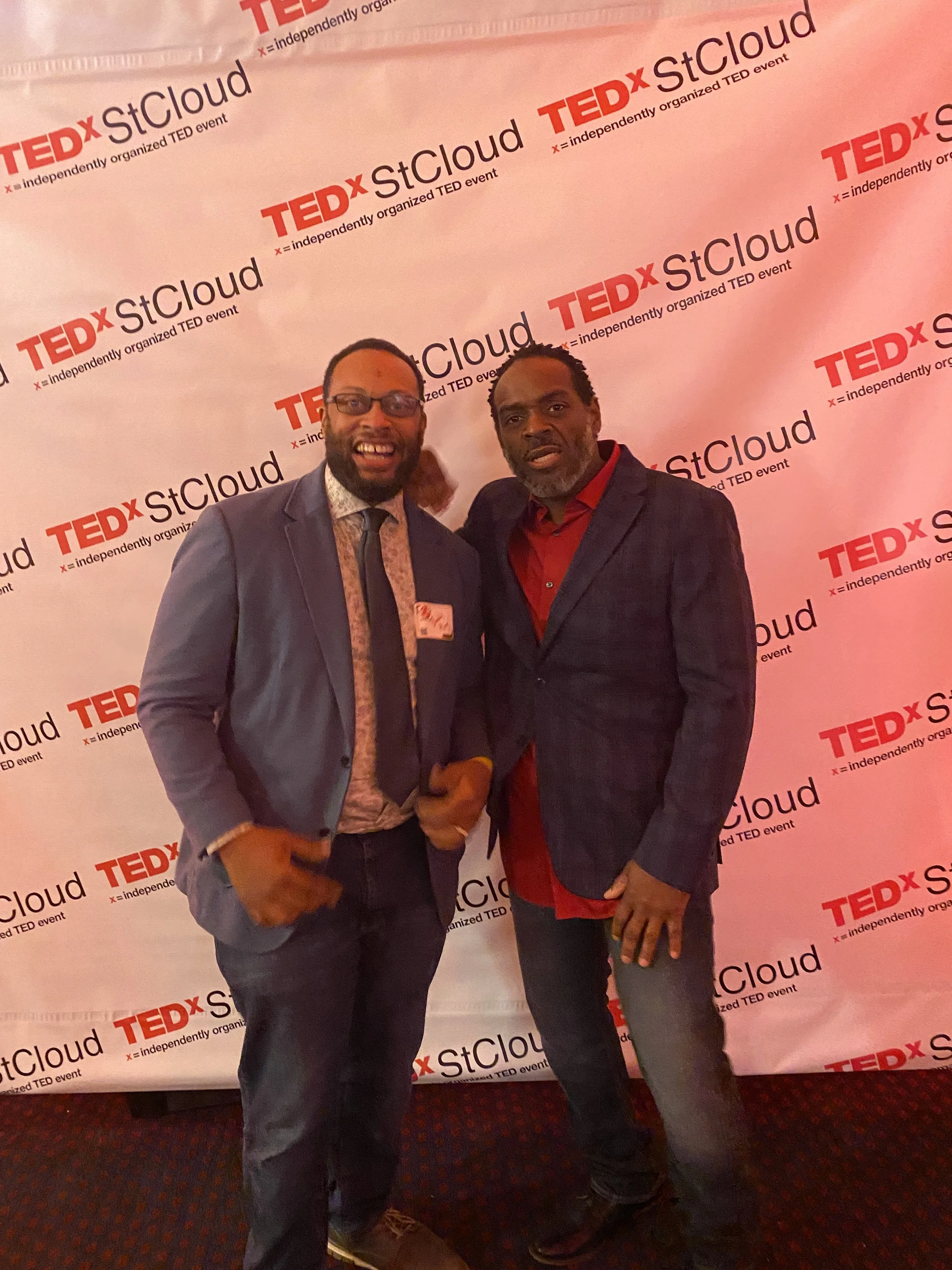 Two men in suits standing in front of a TEDx StCloud backdrop at an event.