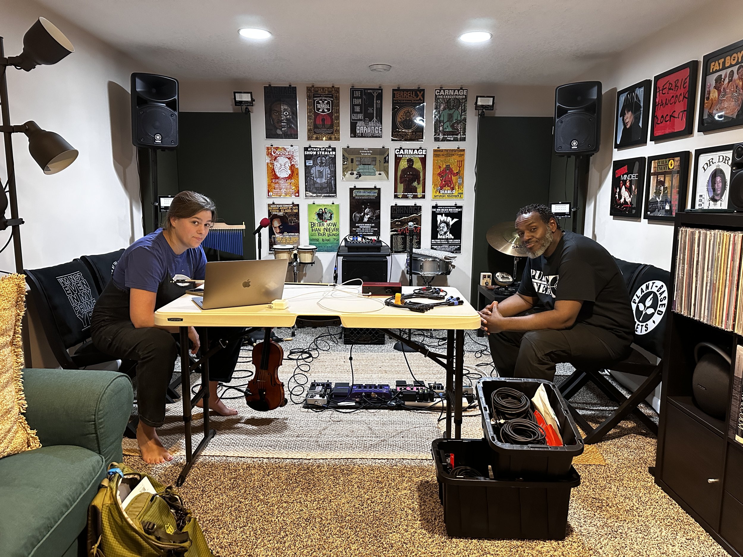 Two people are in a music practice room. One person is sitting at a table with a laptop, and the other is sitting nearby with musical equipment in front of them. The room has walls decorated with musical posters and speakers.