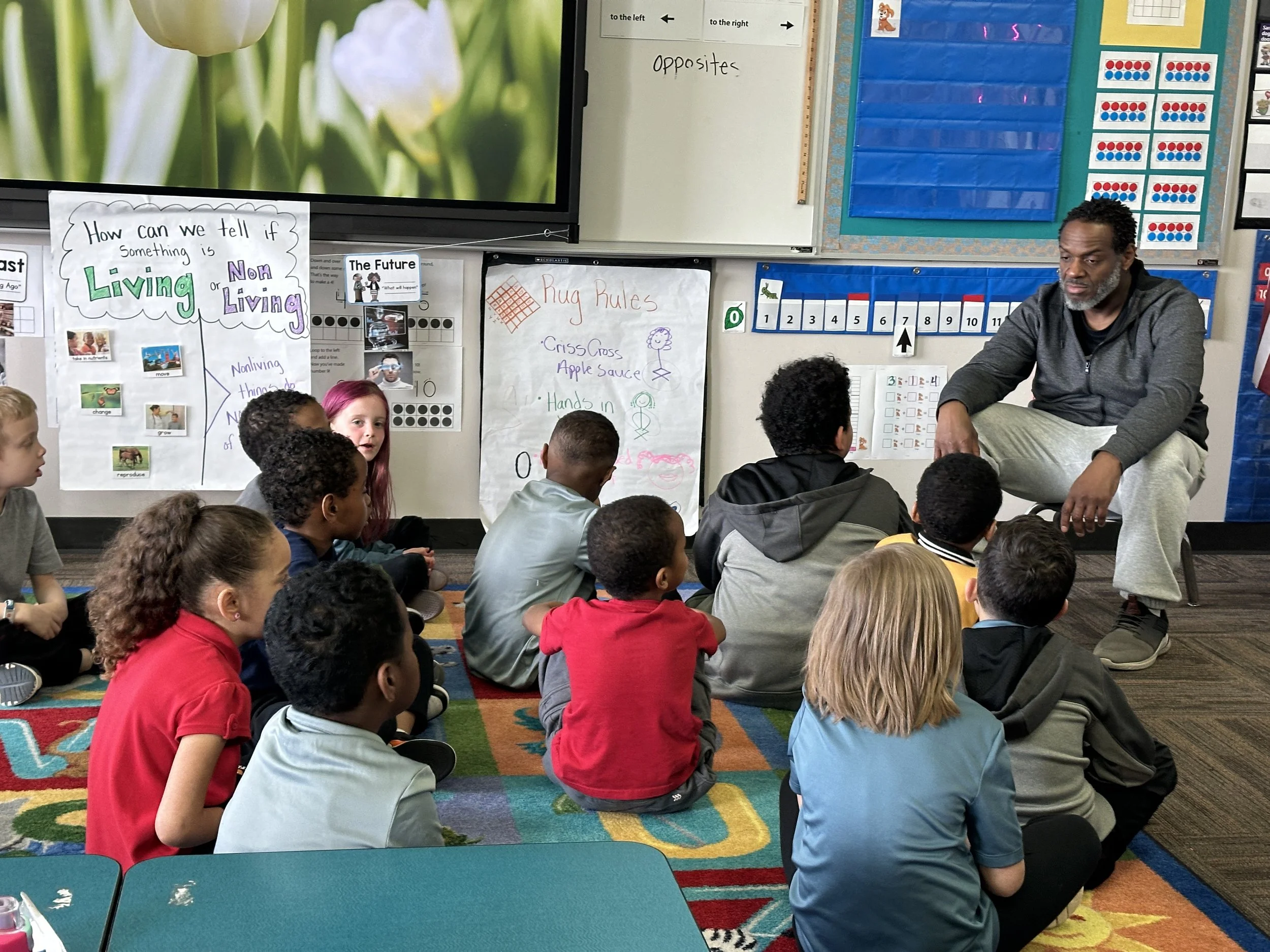A teacher sitting on a chair in front of a classroom of middle school students seated on a colorful carpet. The classroom has educational posters and charts on the walls, including one about living and nonliving things and another about rug rules. Th