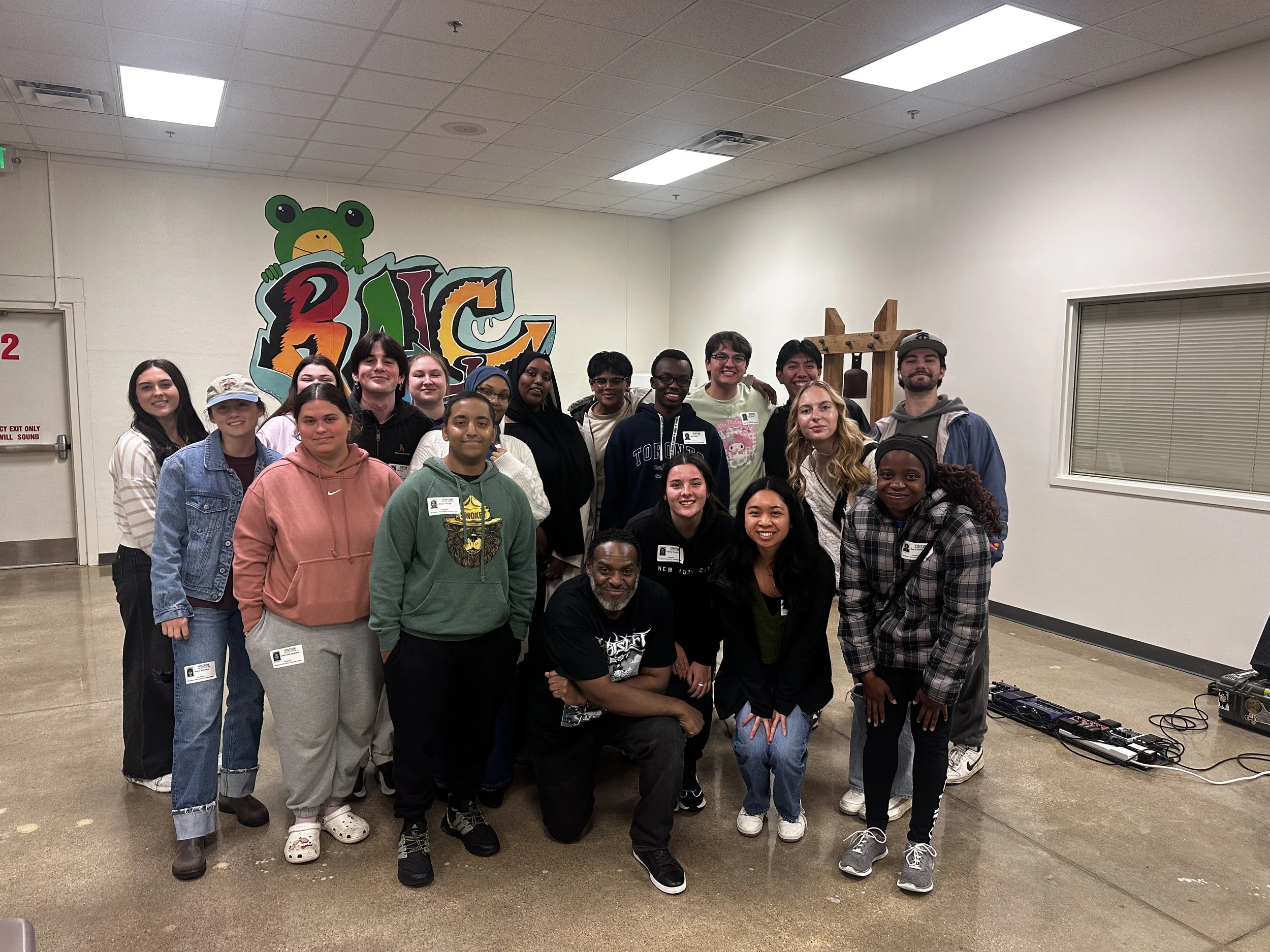 Group of diverse young people gathered indoors in front of a colorful graffiti-style mural with a frog and letters, smiling and posing for a photo.