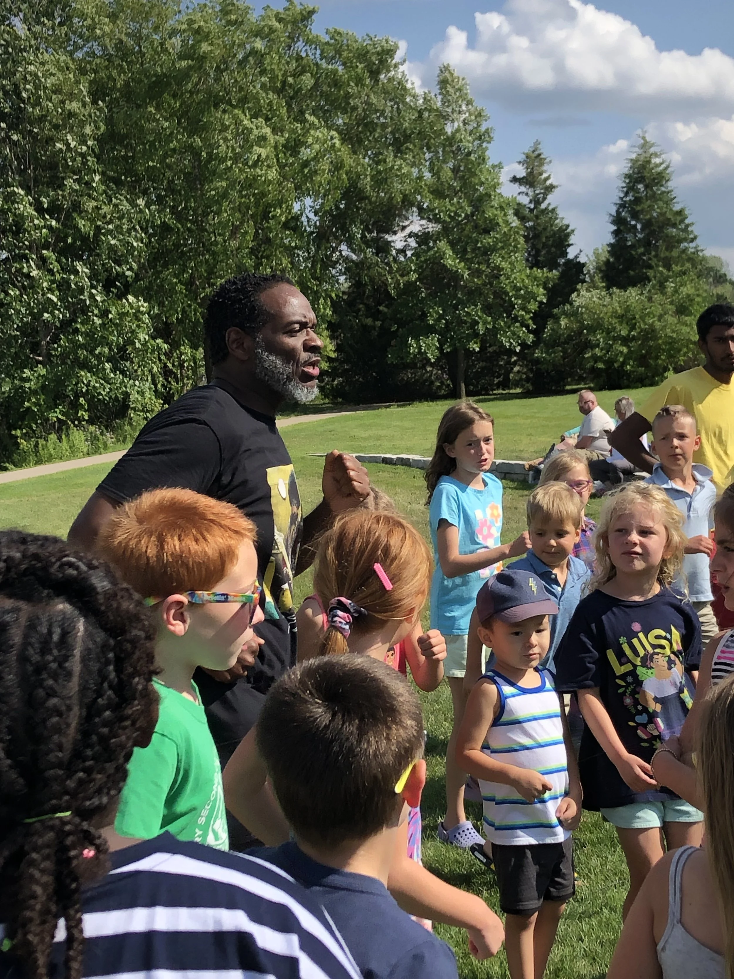 A group of children and adults gathered outdoors on a sunny day with green trees, grass, and a blue sky with clouds in the background. One adult man with a gray beard appears to be speaking or giving instructions, and the children are listening attentively.