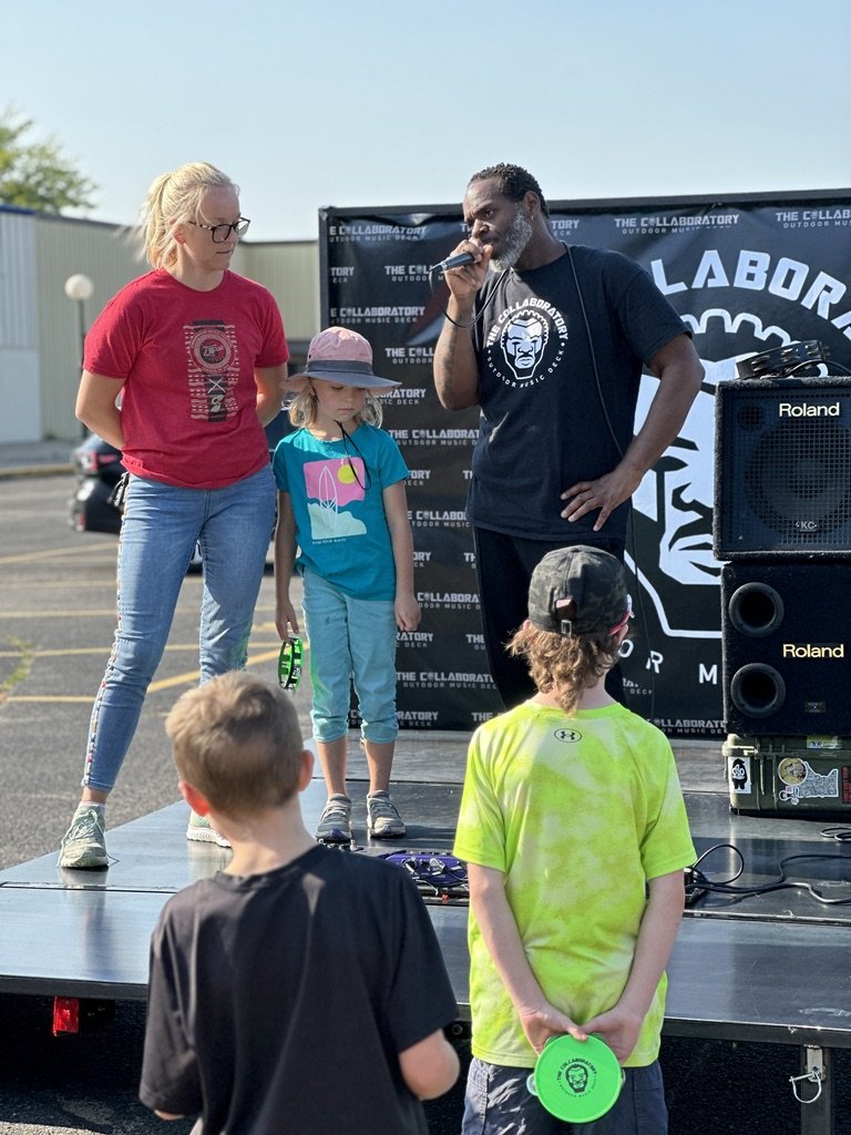 A man with a microphone on a small outdoor stage, flanked by a woman and a girl, is speaking to or performing for three children watching in front. The event is outdoors with a banner that says 'The Collaboratory Outdoor Music Deck.'