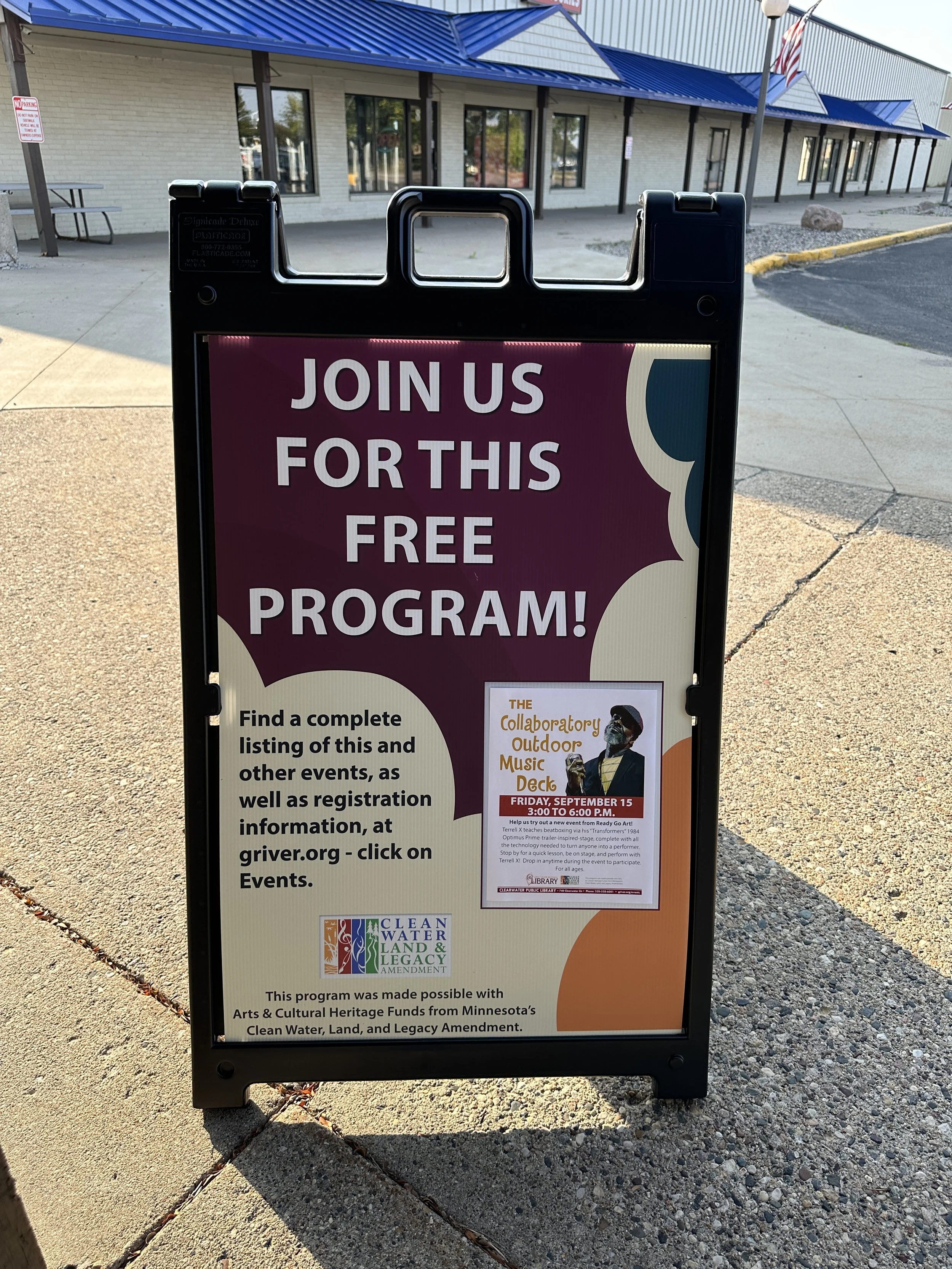 Outdoor sidewalk sign promoting a free event, with details about a music deck event scheduled for September 15 from 3:00 to 6:00 p.m., including a description of the event and registration instructions, with a building in the background and a no parking sign nearby.