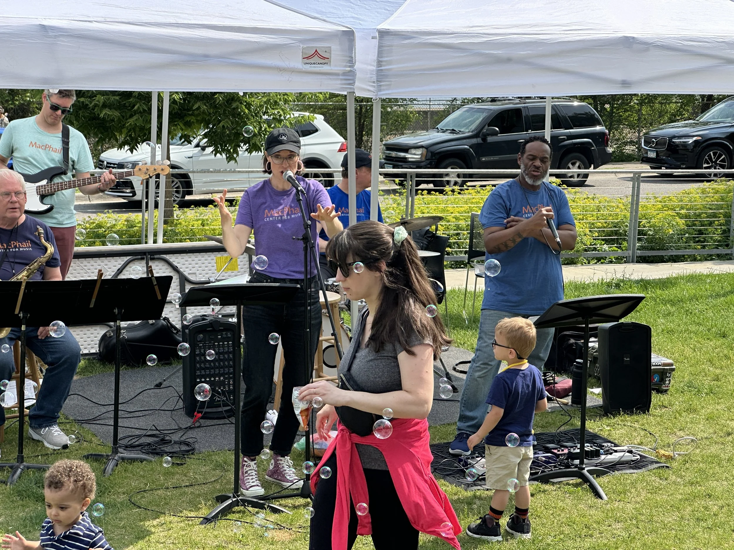 A group of people performing music outdoors under a white canopy, including a woman singing into a microphone, a man playing the guitar, and others playing instruments. Children and audience members are present nearby, with some bubbles floating in t