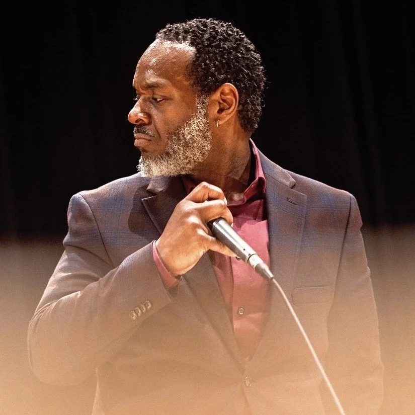 A man with gray and black hair, gray beard, wearing a maroon shirt and suit, holding a microphone, looking to the side against a dark background.