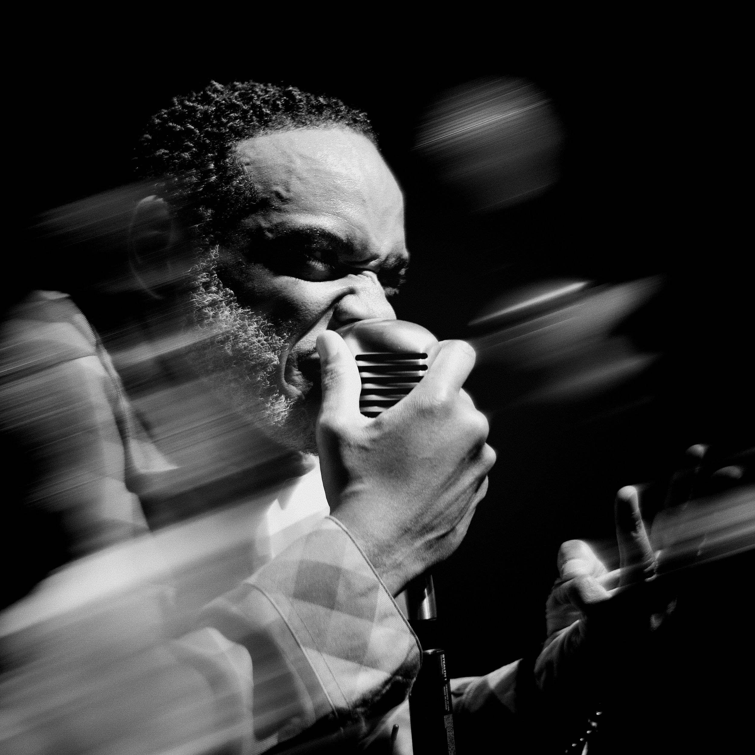 Close-up black and white photograph of a man with curly hair and beard, singing into a microphone with a passionate expression.