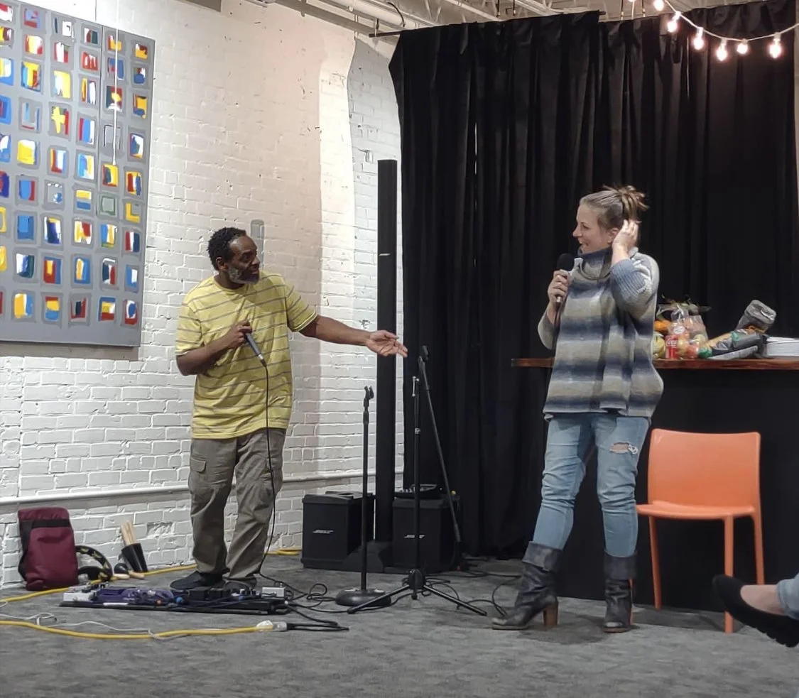 A man and woman are on stage engaging in a comedy or spoken word performance. The man is holding a microphone and gesturing with his hand, while the woman is smiling and holding a microphone, dressed casually in boots and ripped jeans. The stage is s