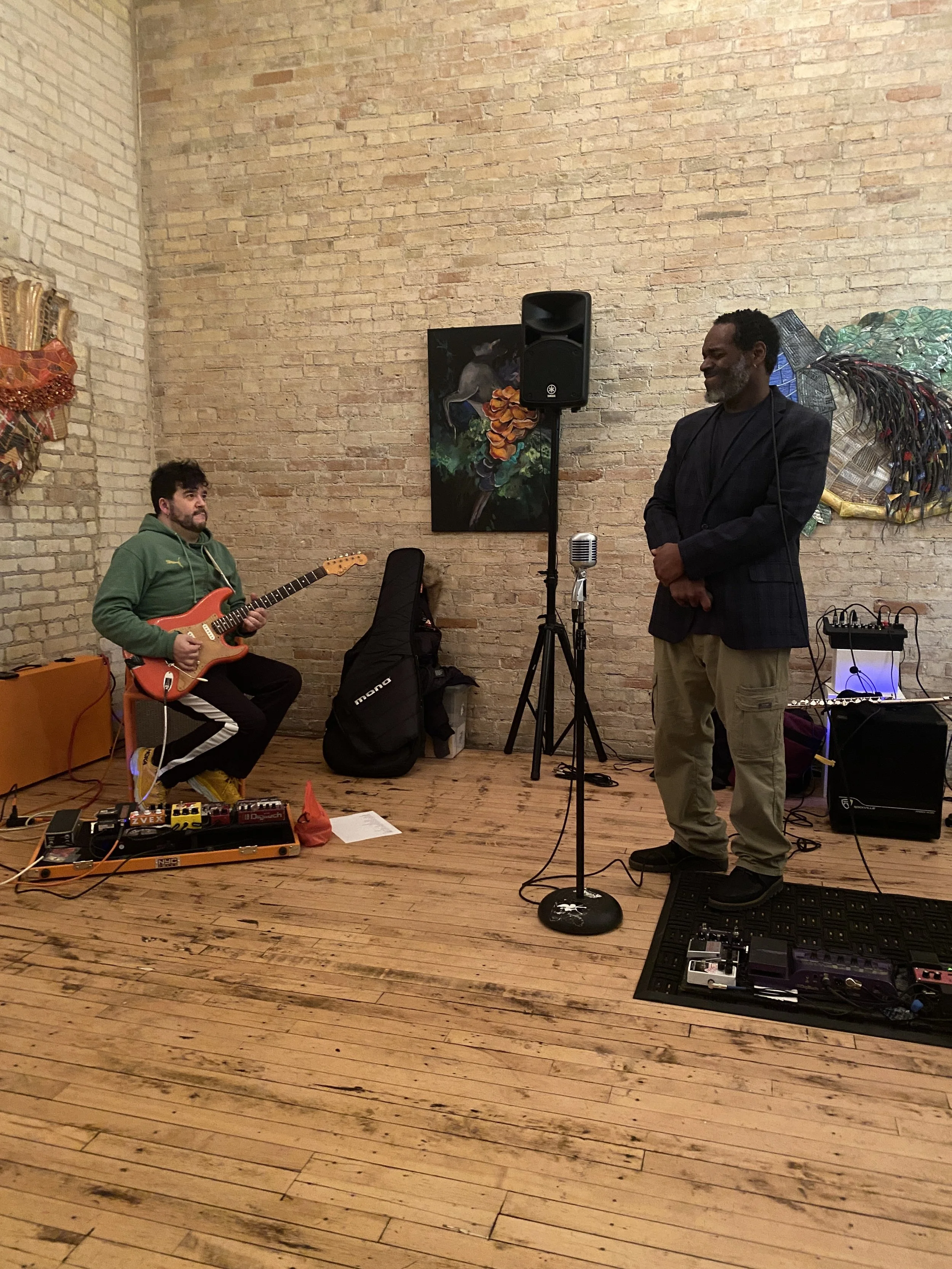 A musician playing electric guitar and a man standing near a microphone in a brick-walled room with artworks and musical equipment.