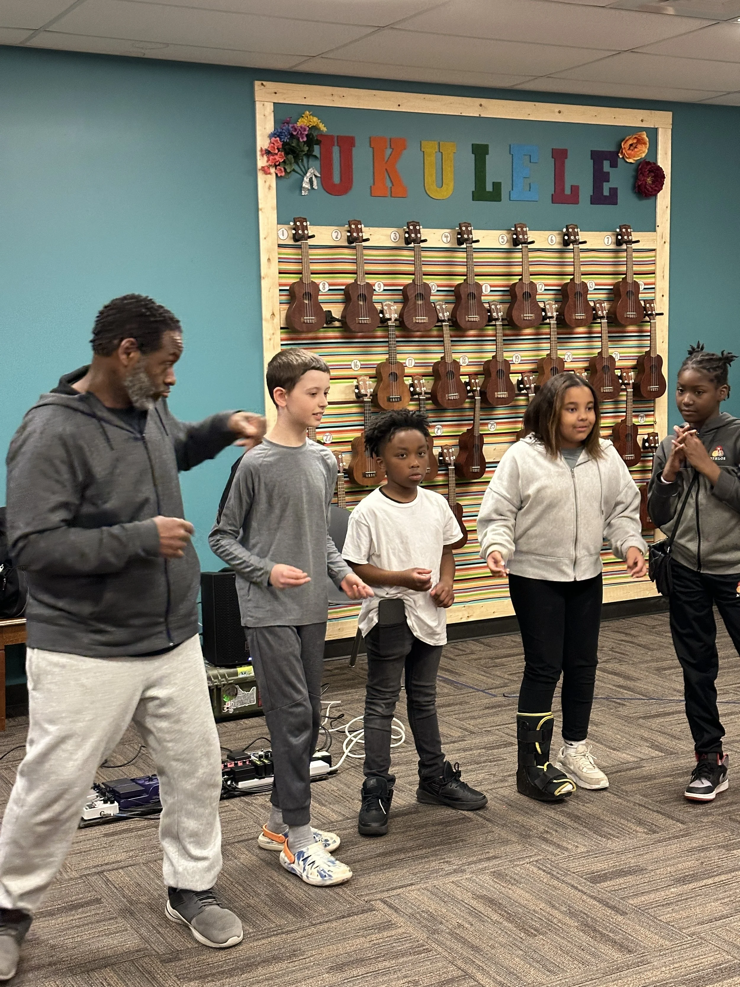 A group of children and an adult standing in a classroom or community center, with ukuleles mounted on a colorful striped backdrop, and the word 'UKULELE' displayed above in multicolored letters.