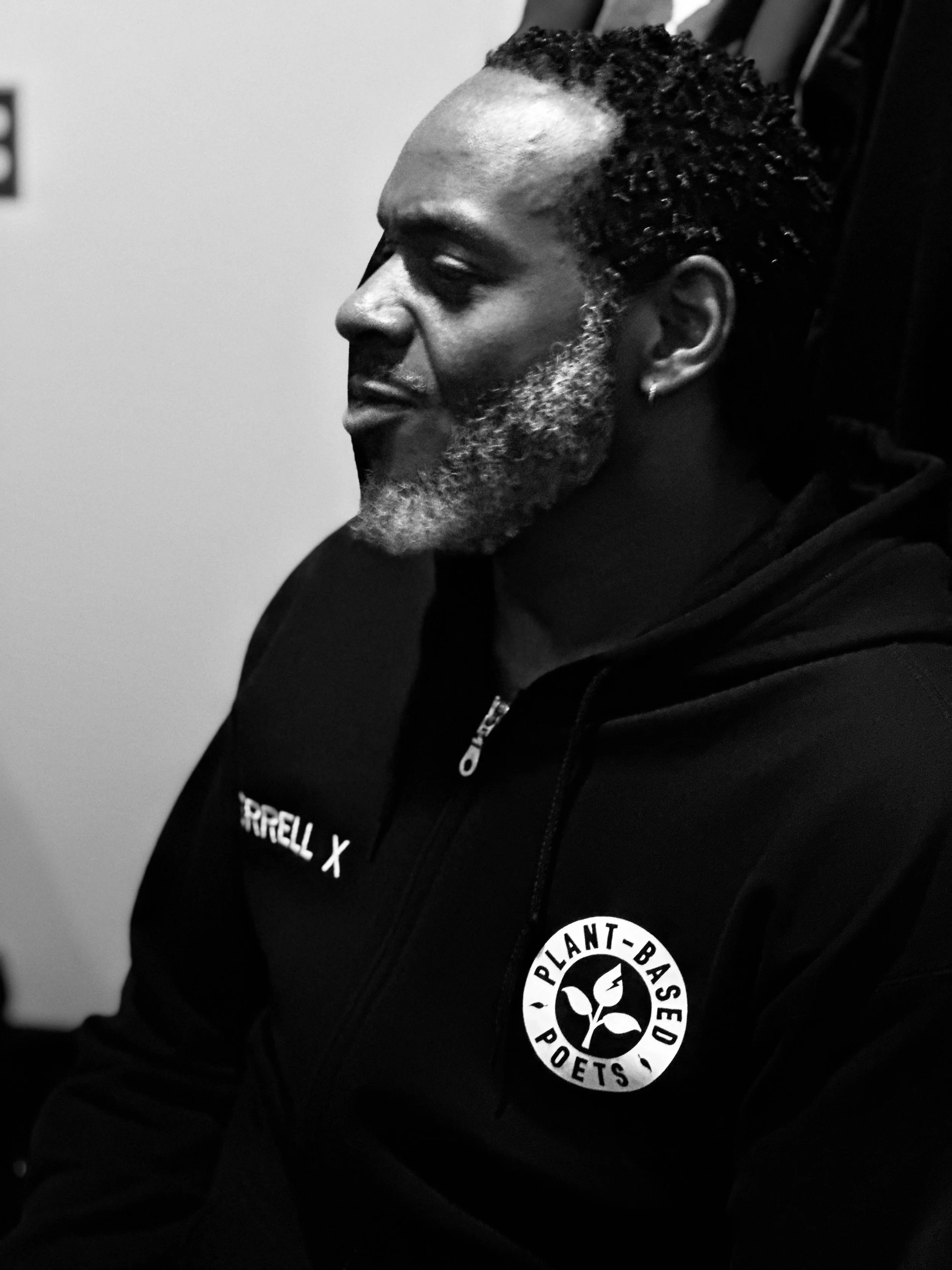 Black and white photograph of a man with braided hair, beard, and earring, wearing a hooded sweatshirt with patches, one saying 'Plant-Based Poets'.