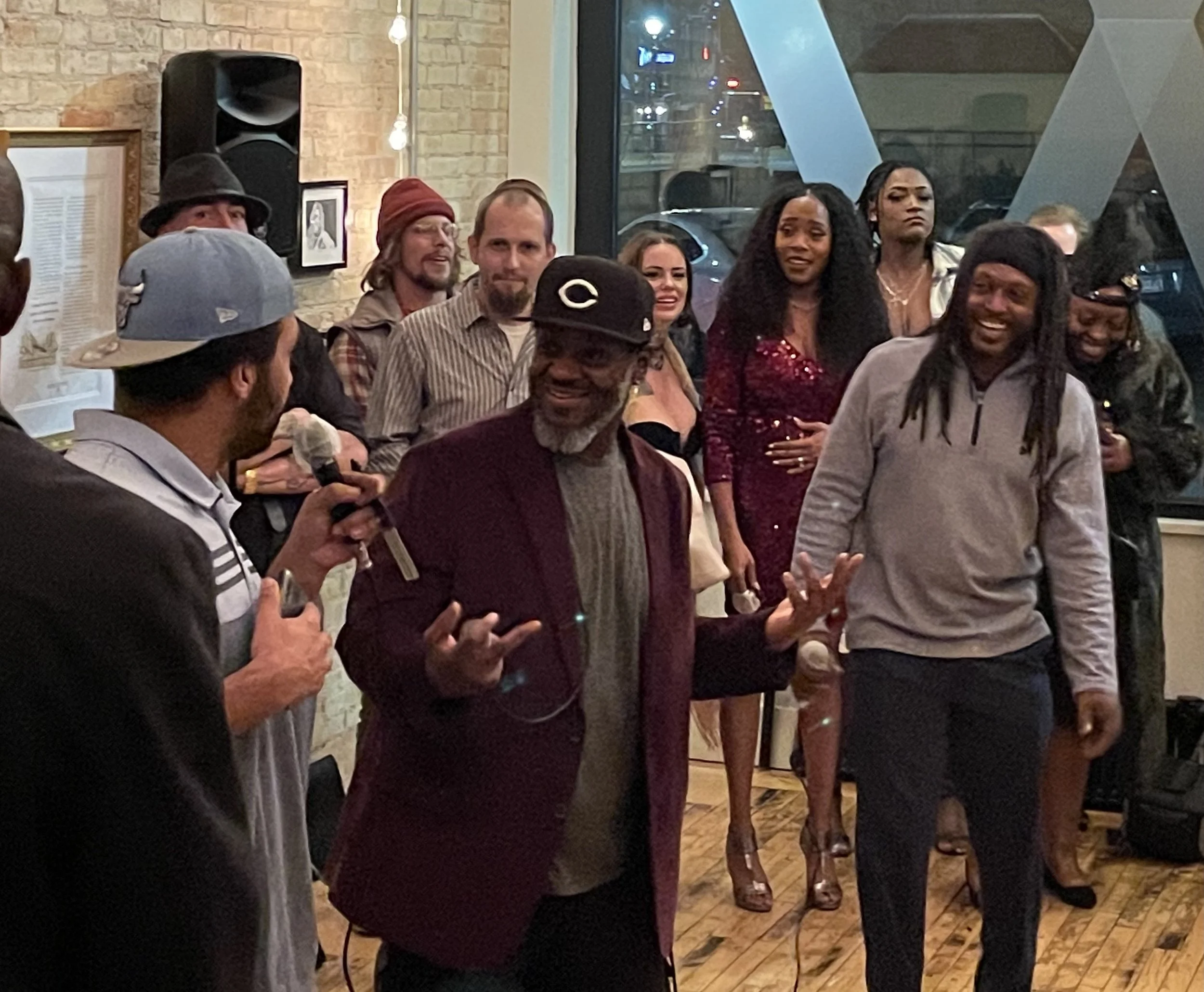 A group of people gathered indoors, some smiling, listening to a man holding a microphone. The room has a brick wall, large windows, and artwork on the walls, indicating a celebratory event.