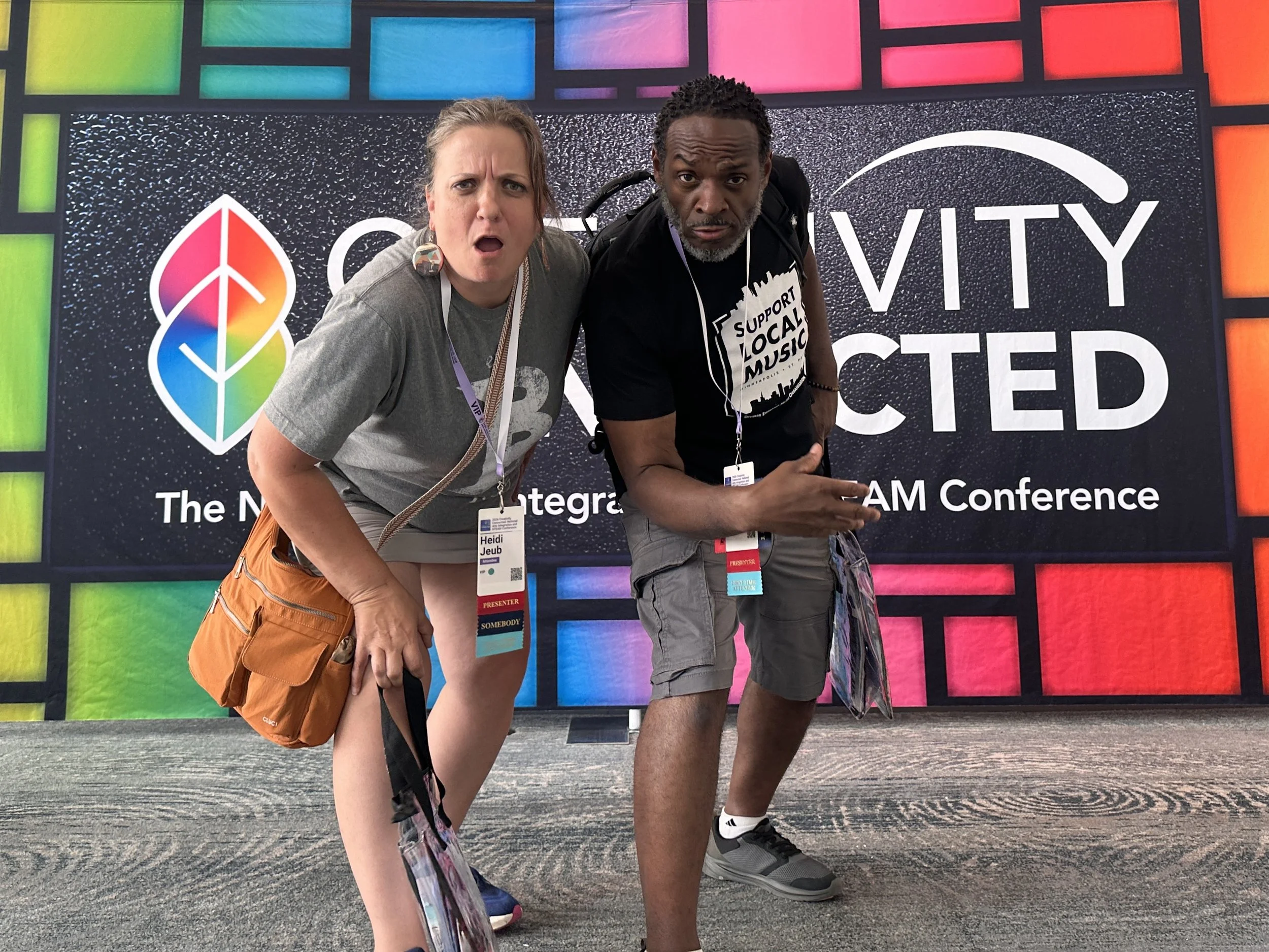 Two people posing in front of a colorful conference backdrop. The woman on the left wears a gray shirt and anorange sachel, and the man on the right wears a black t-shirt with a support local music message. They appear surprised and slightly puzzled.