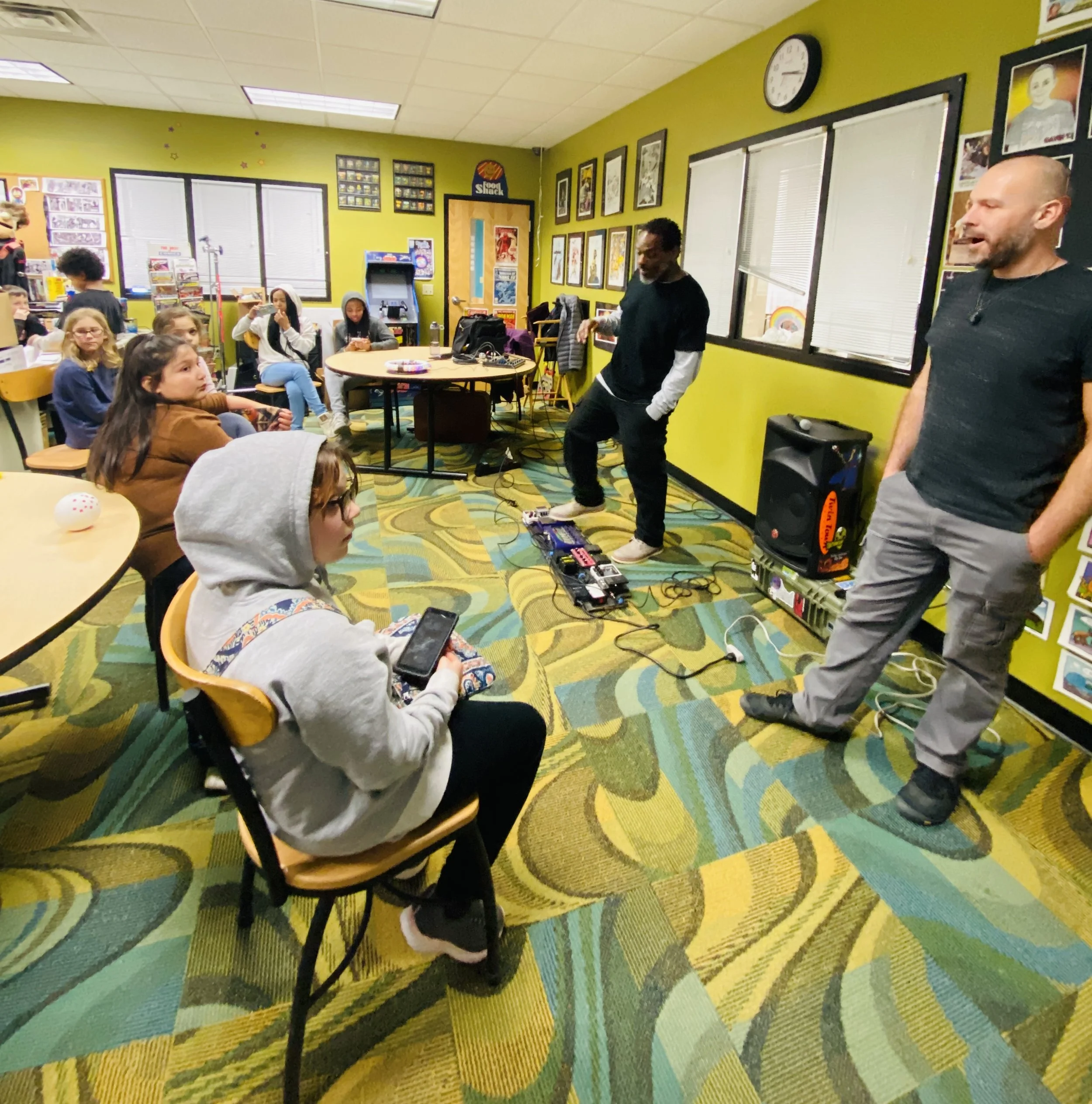 A group of children and two adults in a colorful room with green walls and framed artwork. One child is sitting on a chair, and others are seated or standing, some taking photos or videos. The room has large windows, a table with electronic music equ