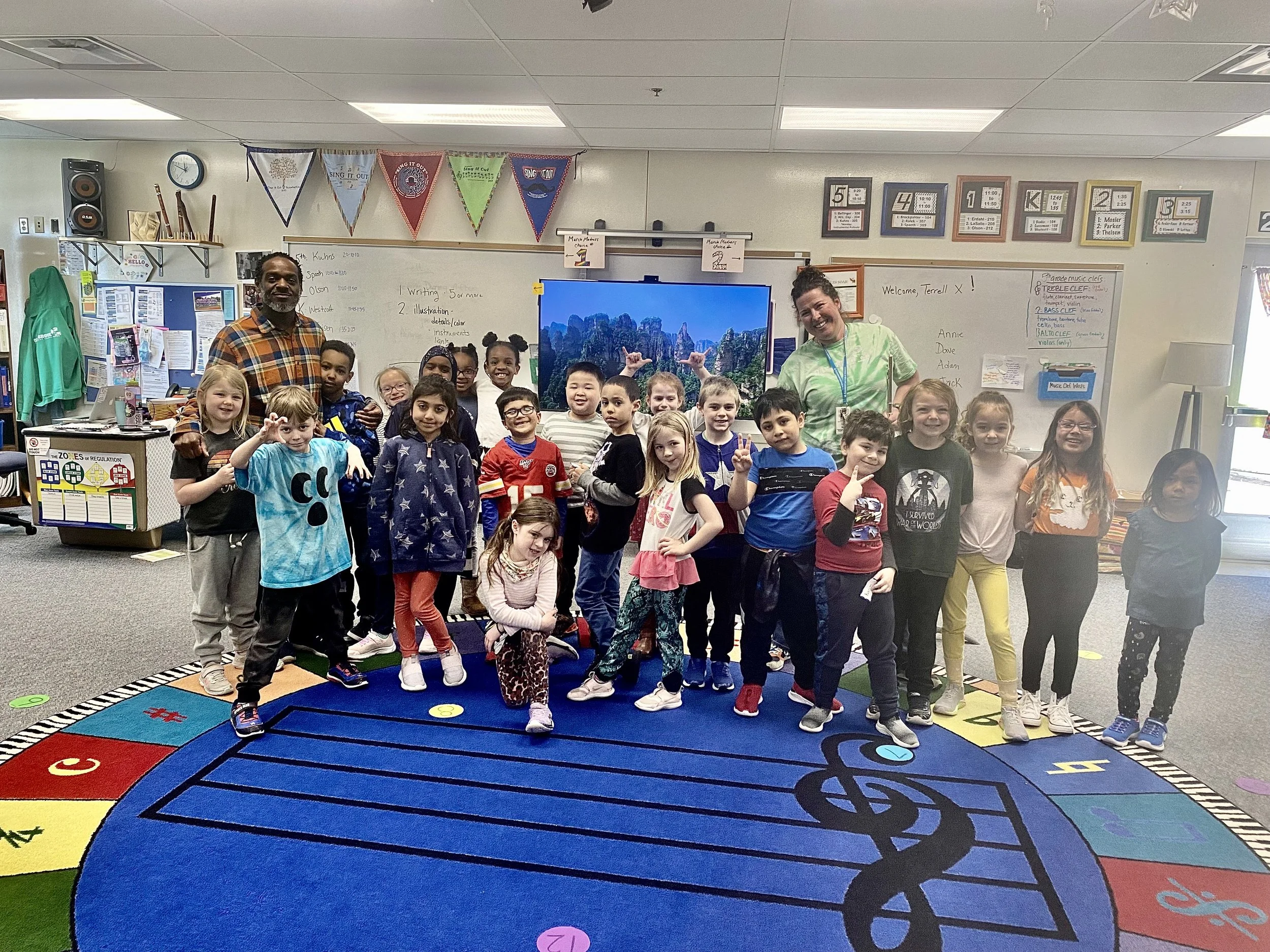 A group of children and two adults in a classroom, standing on a colorful carpet with numbers and letters. The children are smiling and making various hand gestures. The classroom has educational posters, a whiteboard, and banners hanging from the ce