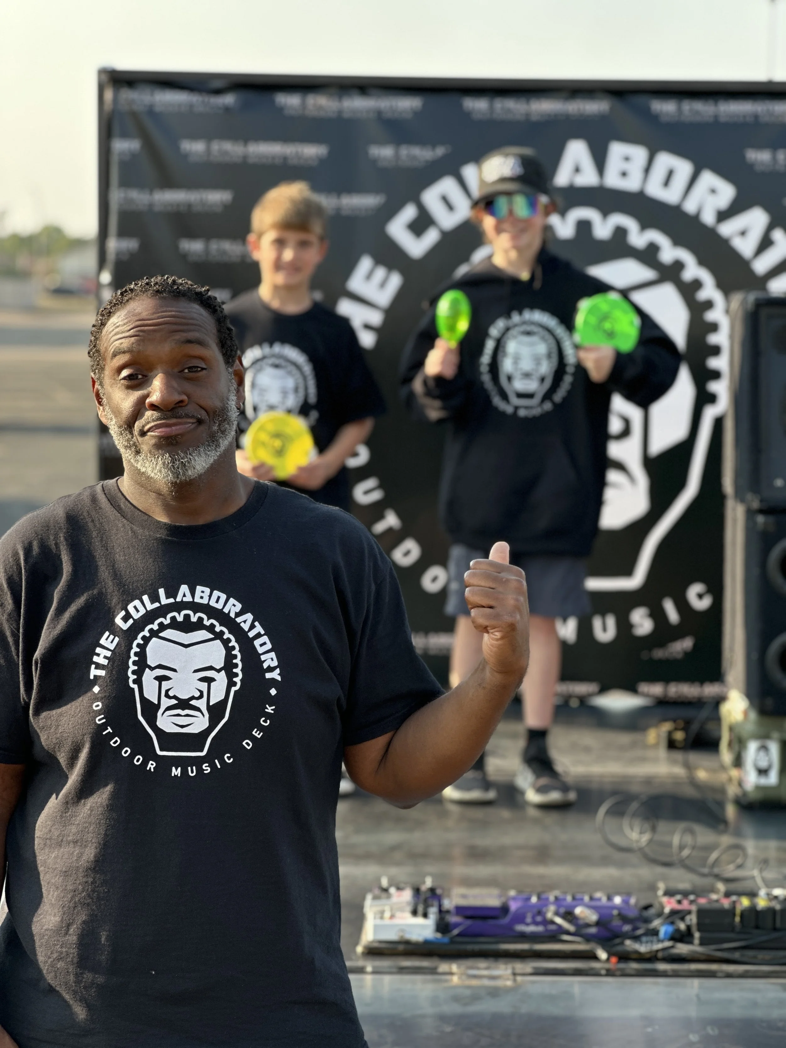 A smiling man with gray beard and hair wearing a black T-shirt with a white logo and text, giving a thumbs-up gesture, with two boys in the background at an outdoor event. The boys are wearing black hoodies with graphic designs, one has sunglasses and a cap, and are holding colored paddles. There is a large banner with partial text and a graphic, and equipment including pedals and cables on the table.