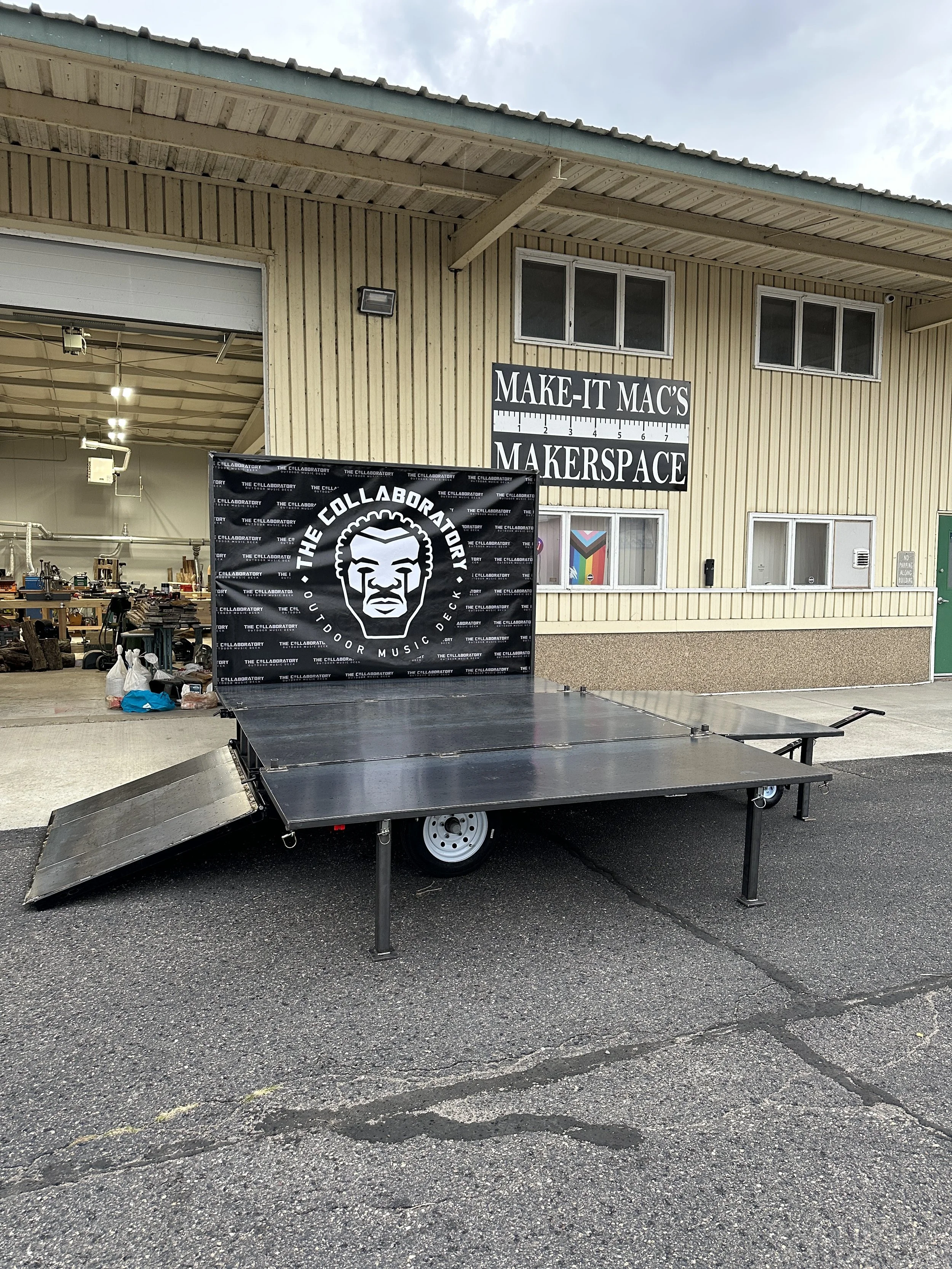 A flatbed trailer with a ramp parked outside a warehouse with signs reading "MAKE-IT MAC'S" and "MAKERSPACE". Inside the warehouse, shelves and equipment are visible, with some bags and items on the ground outside.