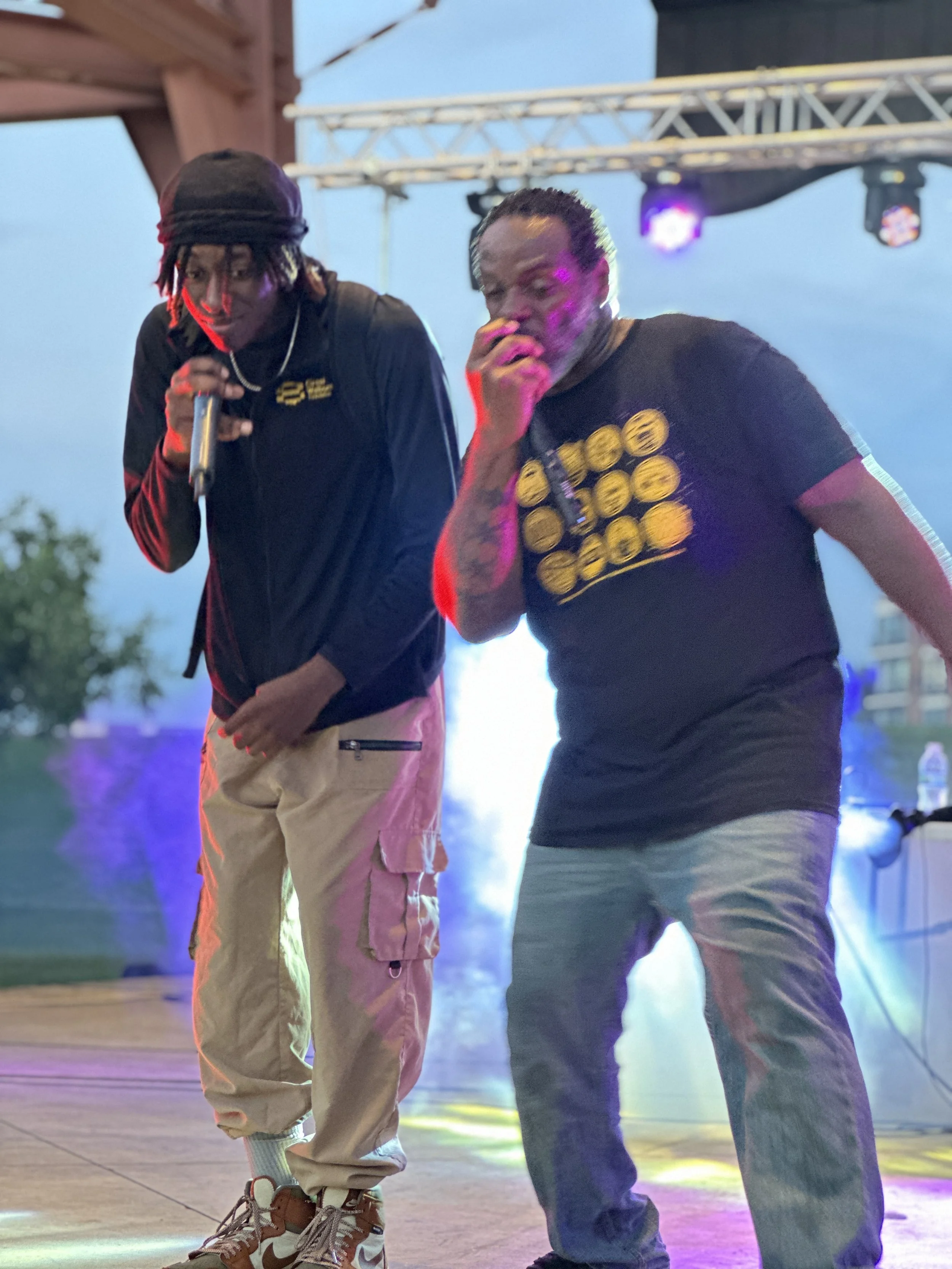 Two young men dance together on an outdoor stage at dusk, with colorful stage lights illuminating the scene. One is wearing a black hoodie, khaki pants, and rollerblades, while the other is dressed in a black T-shirt with yellow graphics and blue jea