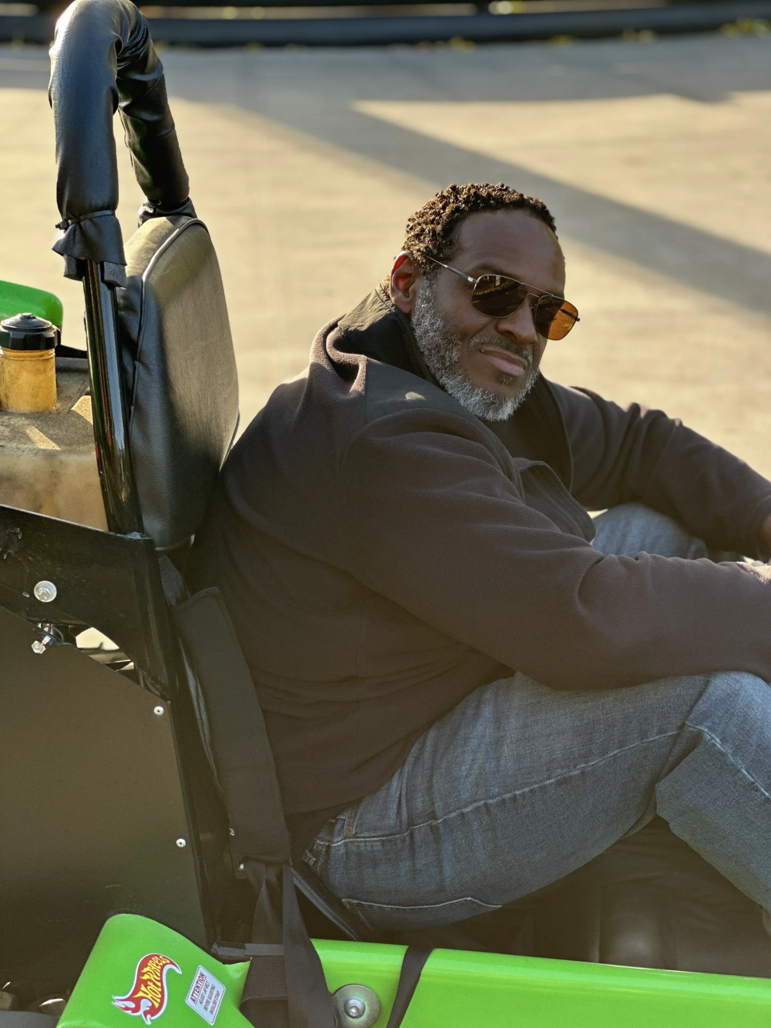 A man with glasses and a beard sitting on a green skateboard, wearing a black hoodie and gray jeans, with a black backpack behind him, outdoors on a sunny day.
