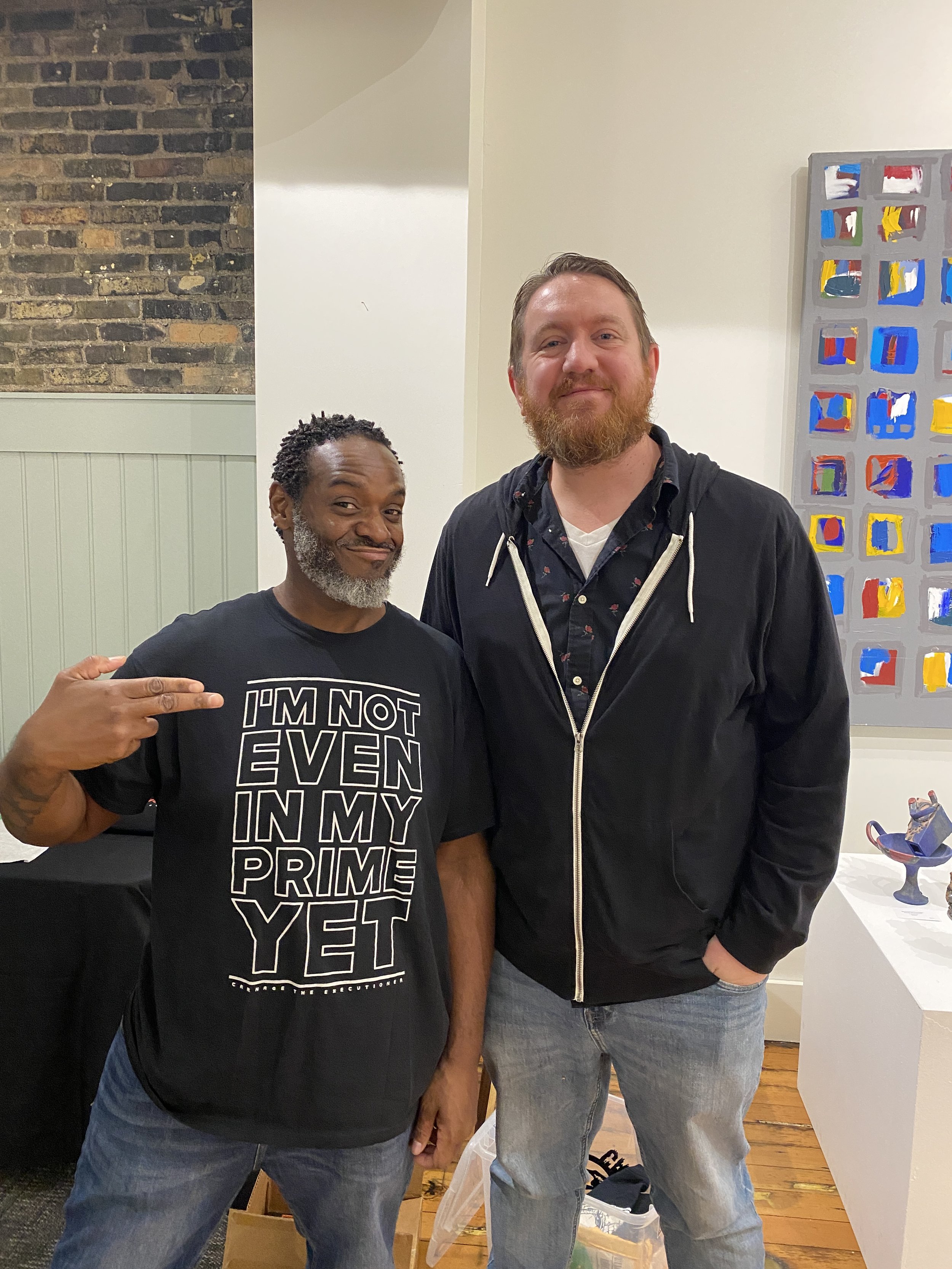 Two men standing together and smiling in an indoor setting, with one man pointing to himself, and a colorful abstract painting on the wall to the right.