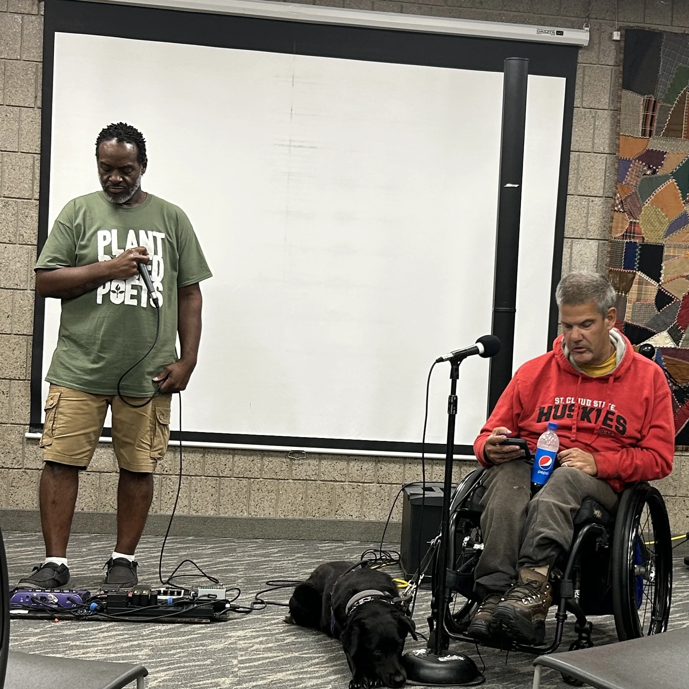A man with dreadlocks wearing a green T-shirt and khaki shorts holding a microphone, a seated man in a wheelchair wearing a red hoodie, and a dog in front of two men, one standing and one sitting, in a room with a whiteboard and patterned carpet.