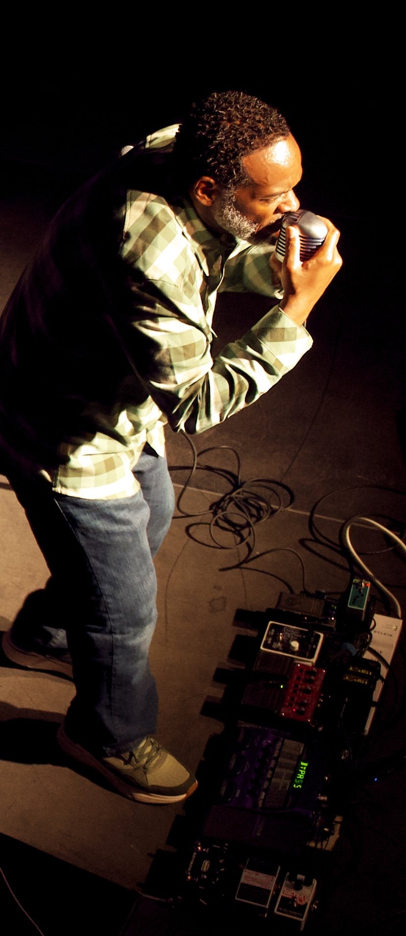 A man singing into a microphone on a dark stage with musical equipment on the floor.