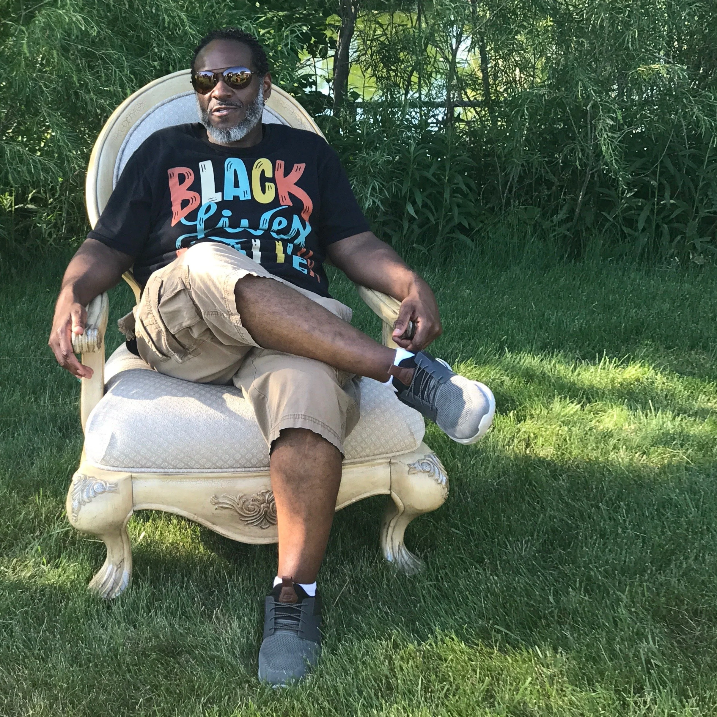 A man with gray hair and beard wearing sunglasses, a black t-shirt with colorful text, and khaki shorts sitting in a vintage armchair outdoors on grass with greenery in the background.